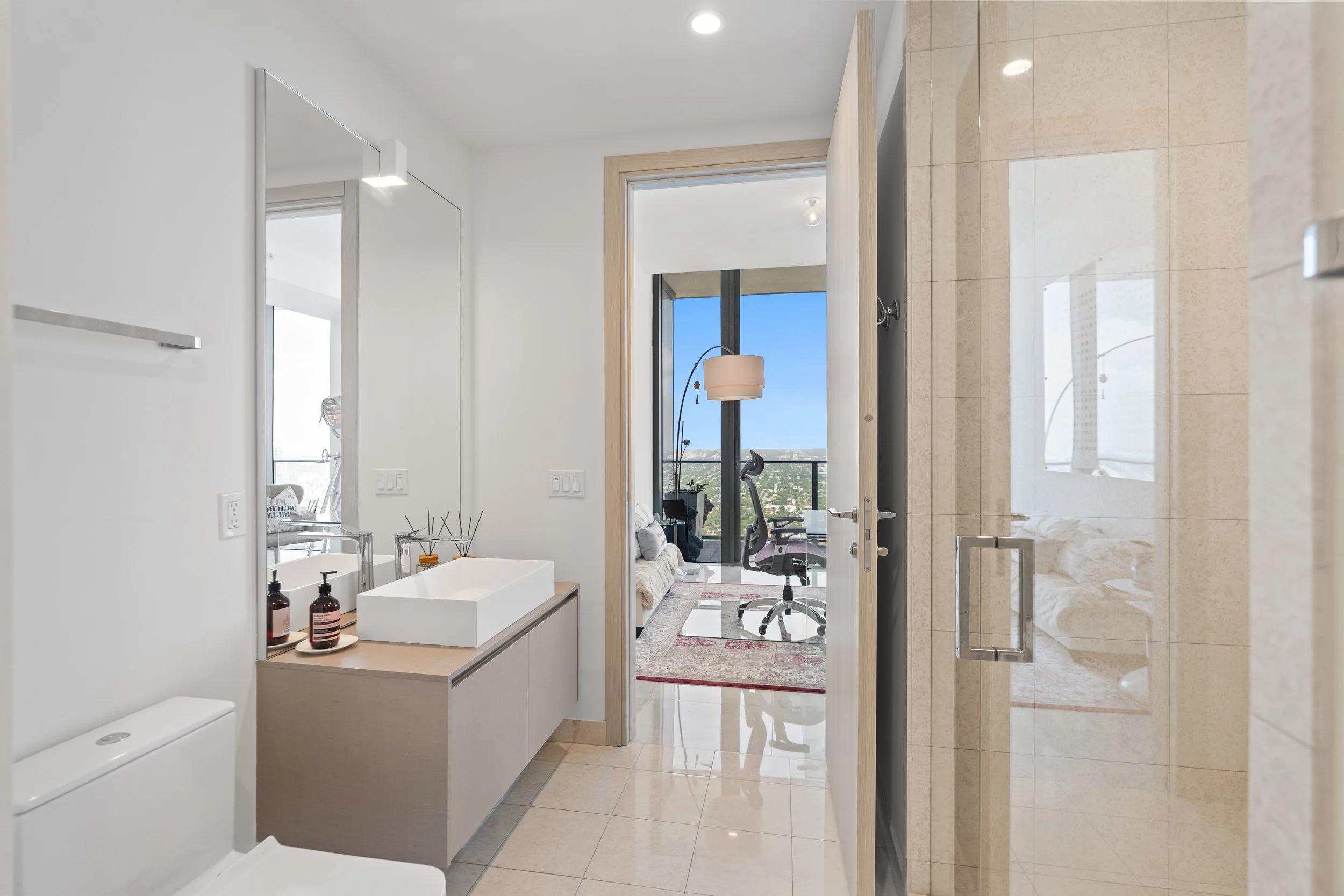 Modern bathroom with a vanity, sink, bottles, and a mirror, leading to a living area with a chair, floor lamp, and a window showing a city view.