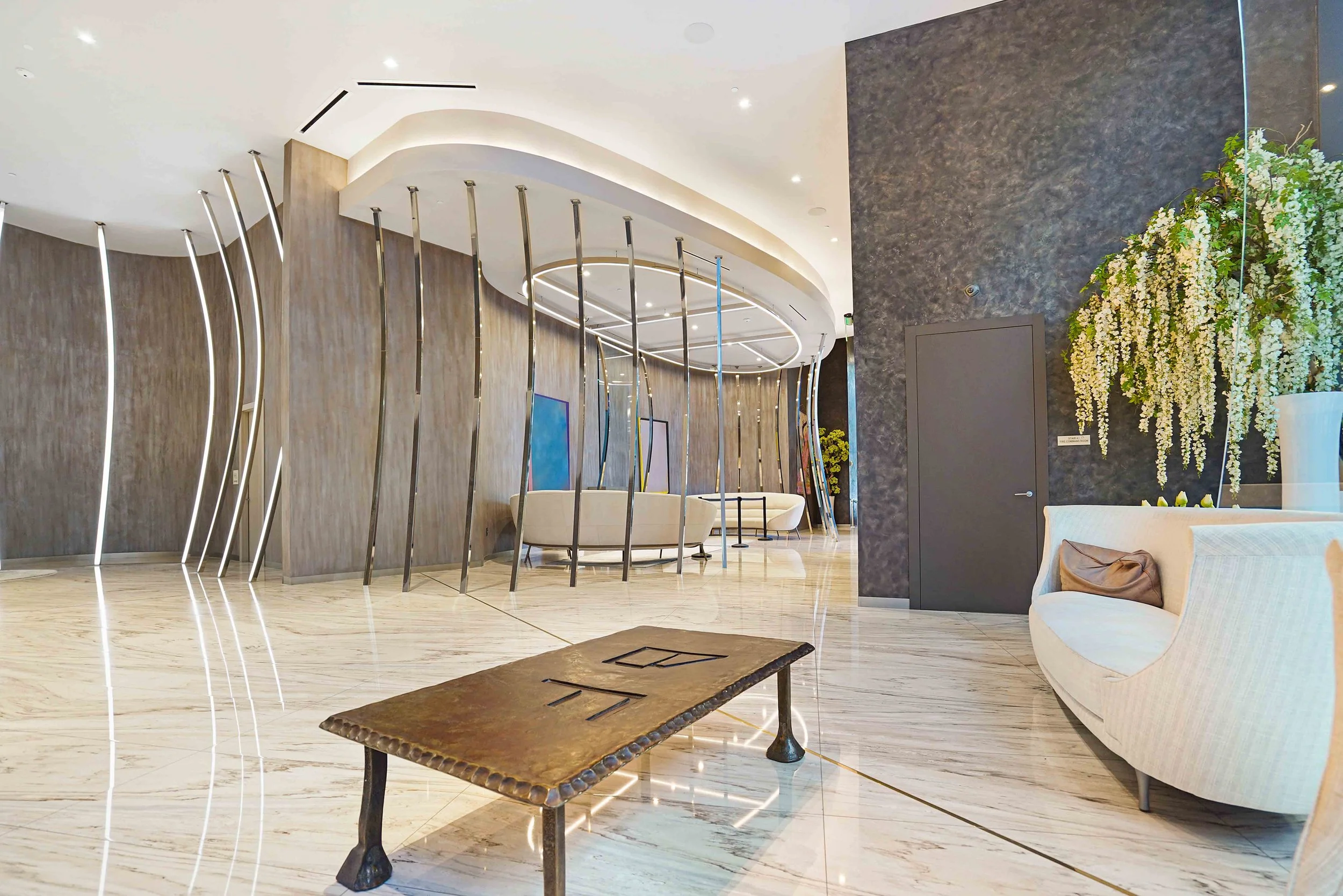Modern hotel lobby with curved white ceiling, decorative metal divider, light-colored marble flooring, large floral arrangement, and seating area with white furniture.