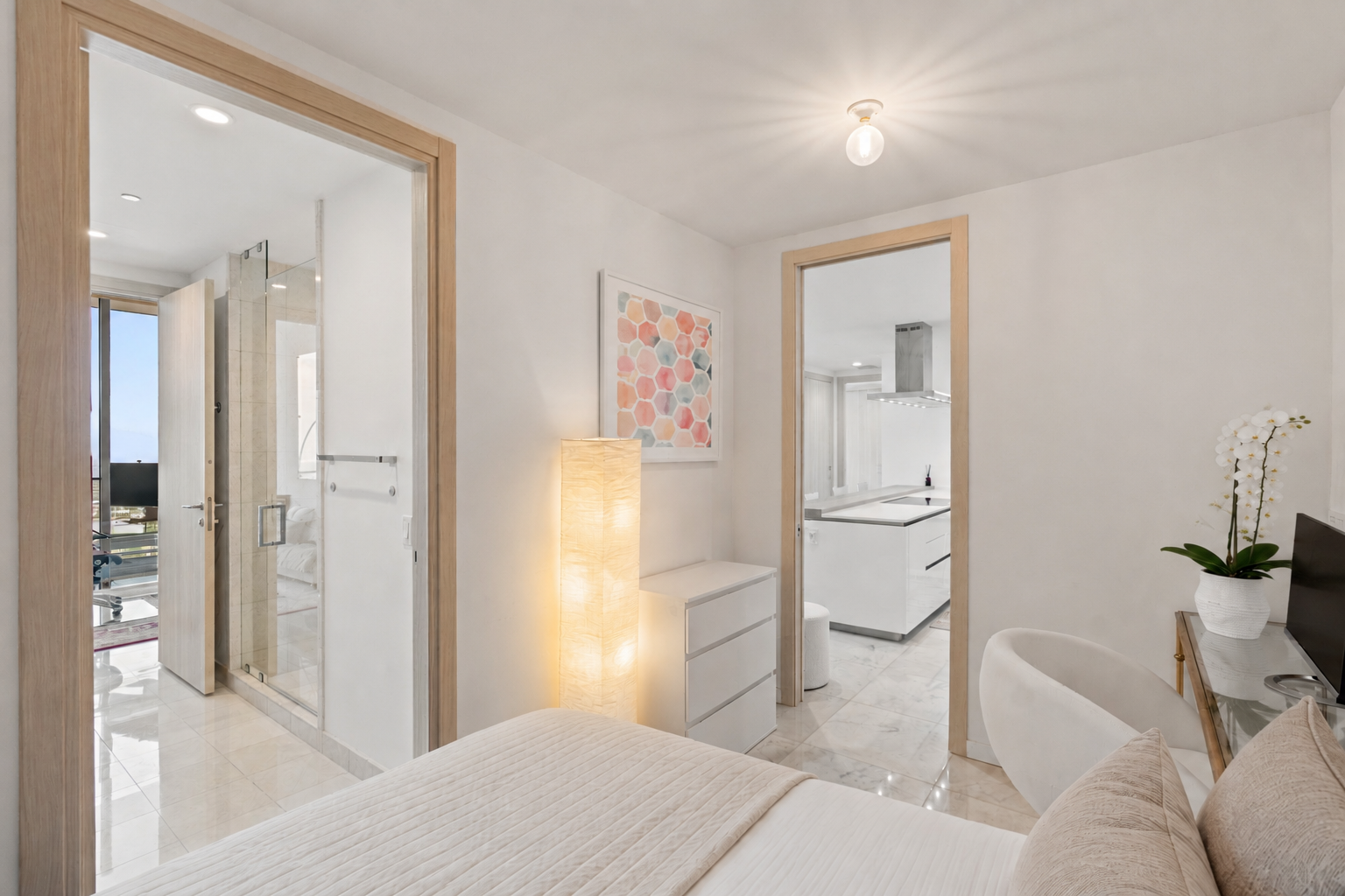 A bedroom with an open door leading to a bathroom and a kitchen area, featuring neutral colors, a white dresser, a floor lamp, an orchid plant, and a television.
