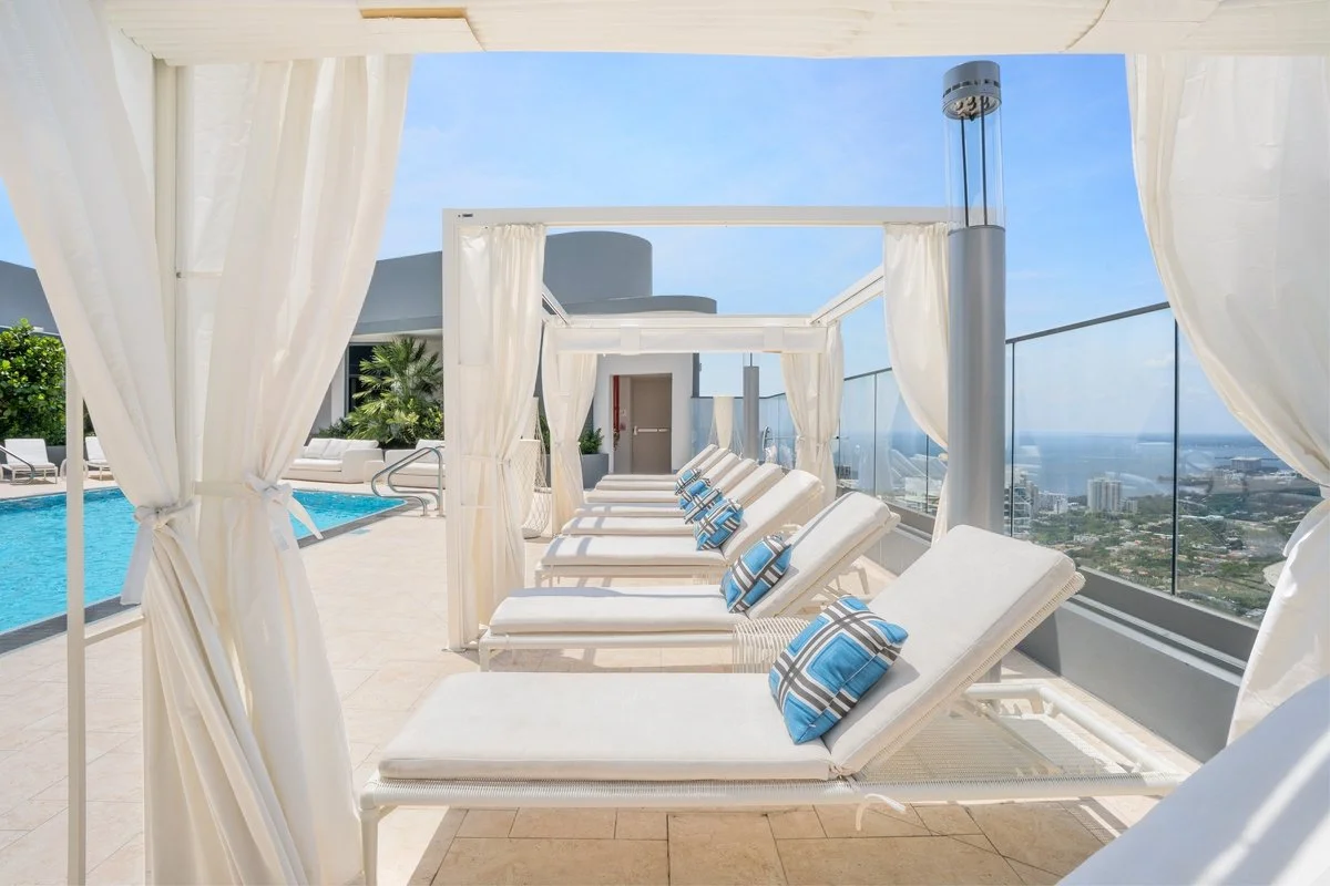 Poolside cabanas with white curtains, lounge chairs with blue and white striped pillows, overlooking a cityscape and ocean on a sunny day.