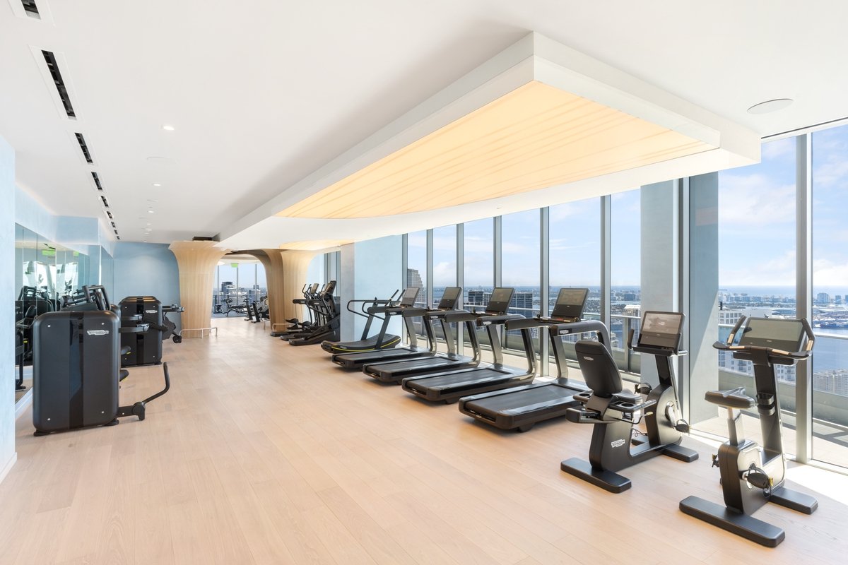Modern gym with treadmills, exercise bikes, and workout equipment near large windows with city view.