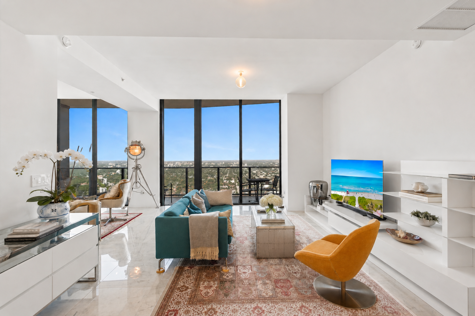 Modern living room with white walls, large windows, a teal sofa with beige pillows, yellow and beige armchairs, a white media console, a flat-screen TV displaying a beach scene, and decor including flowers, books, and a vintage spotlight, with a balcony visible outside.