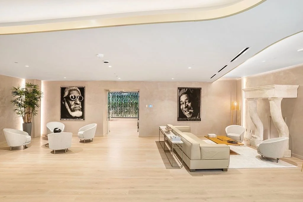 Spacious modern living room with beige walls, white chairs, a white sectional sofa, large photographs of jazz musicians and a potted plant, light wood floors, fireplace with sculpted legs, and a minimalist decor.