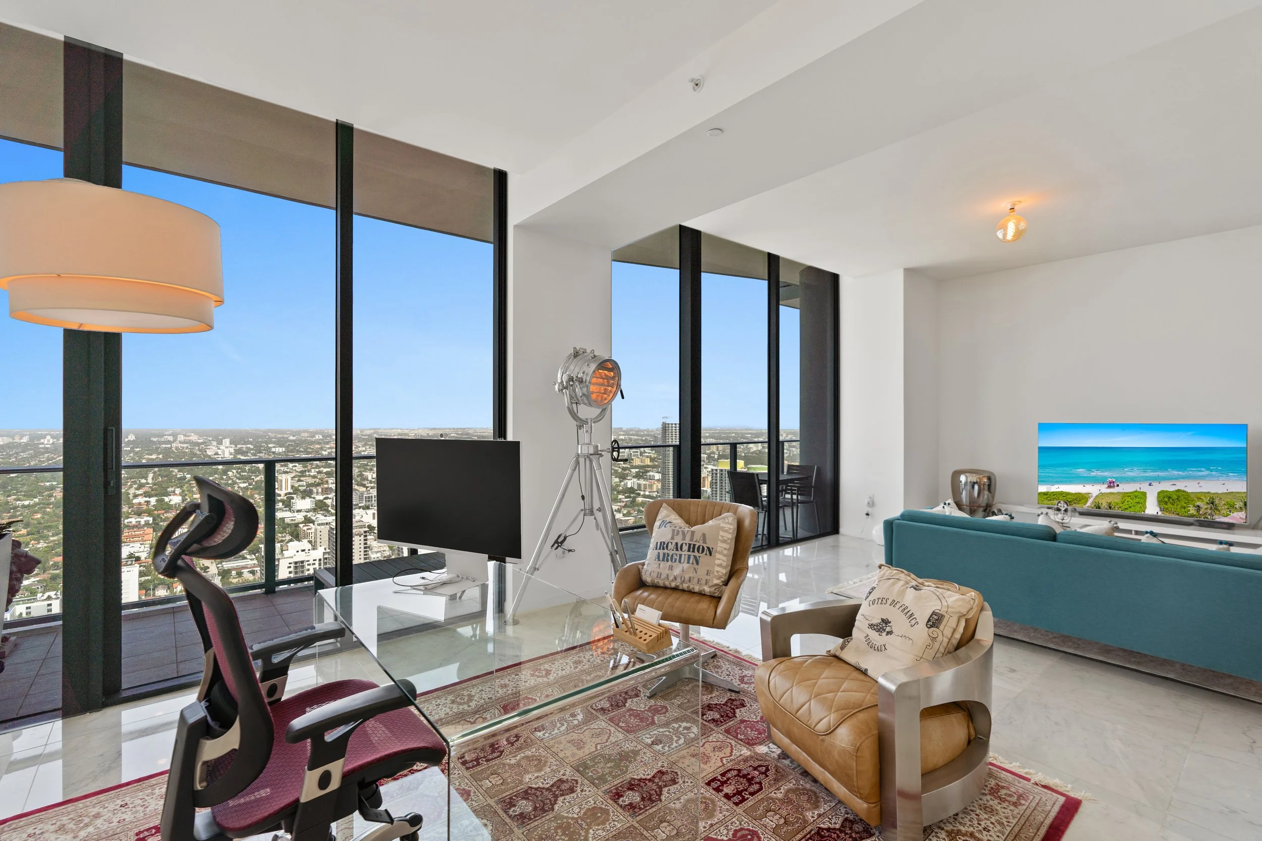 Interior of a modern high-rise apartment with large floor-to-ceiling windows showing a cityscape. The room features a glass dining table surrounded by chairs, a beige armchair, a teal sofa, a flat-screen TV, and various decorative items. There is a l