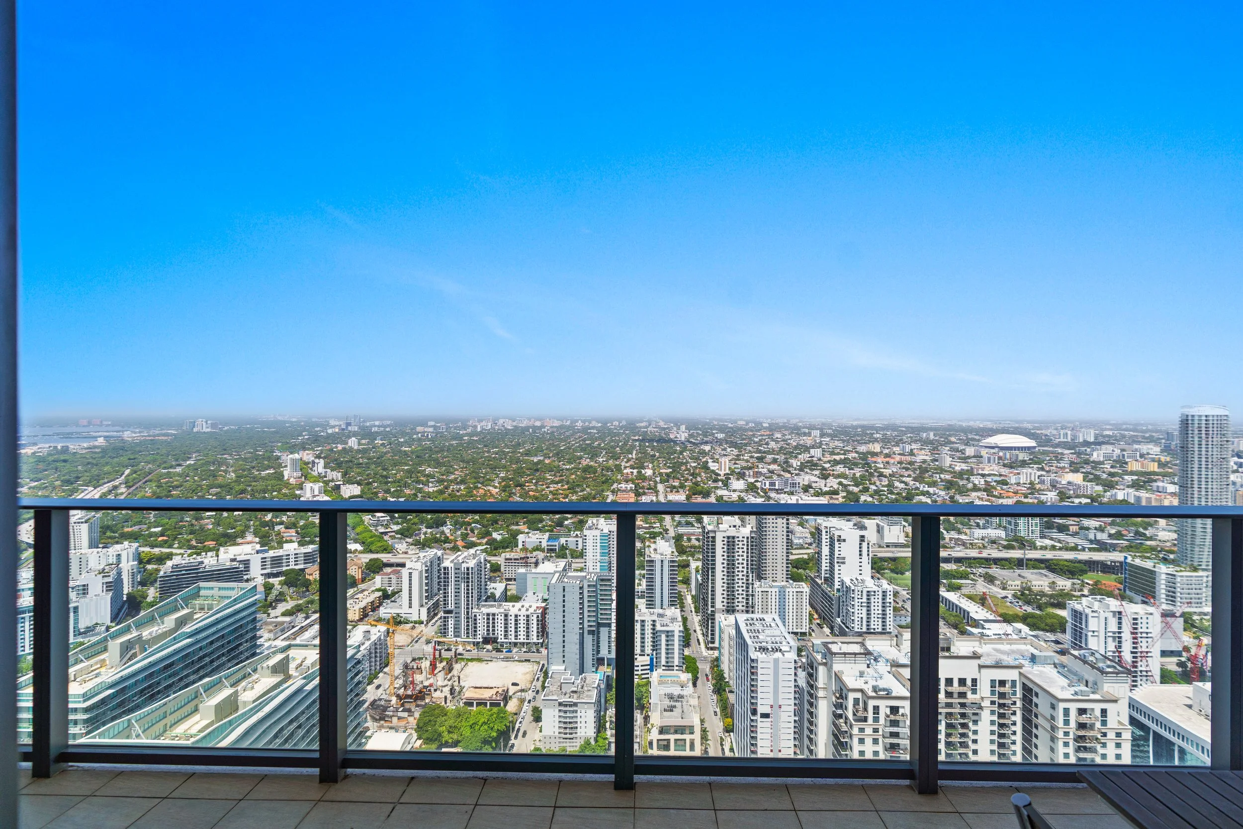 View from a high-rise balcony showing a cityscape with tall buildings, residential areas, and green spaces under a clear blue sky.