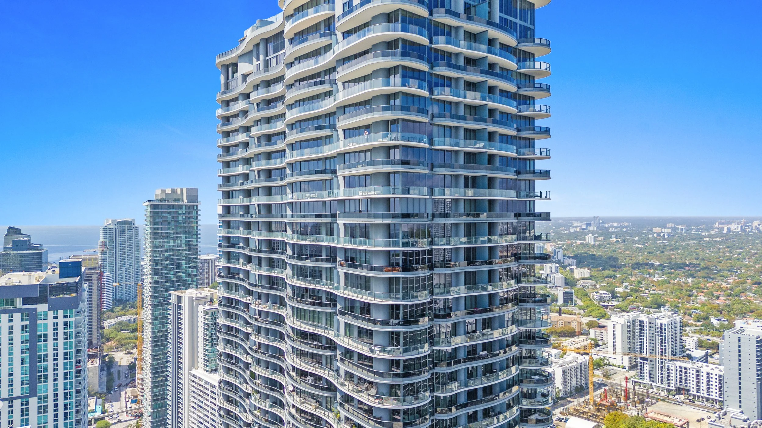 A tall modern skyscraper with curved balconies and glass facade in an urban cityscape with other high-rise buildings under a clear blue sky.