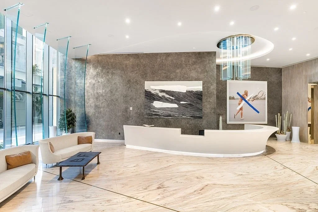 Modern hotel lobby with curved white reception desk, abstract artwork on gray walls, large windows with natural light, and stylish seating area with white couches and a black coffee table.