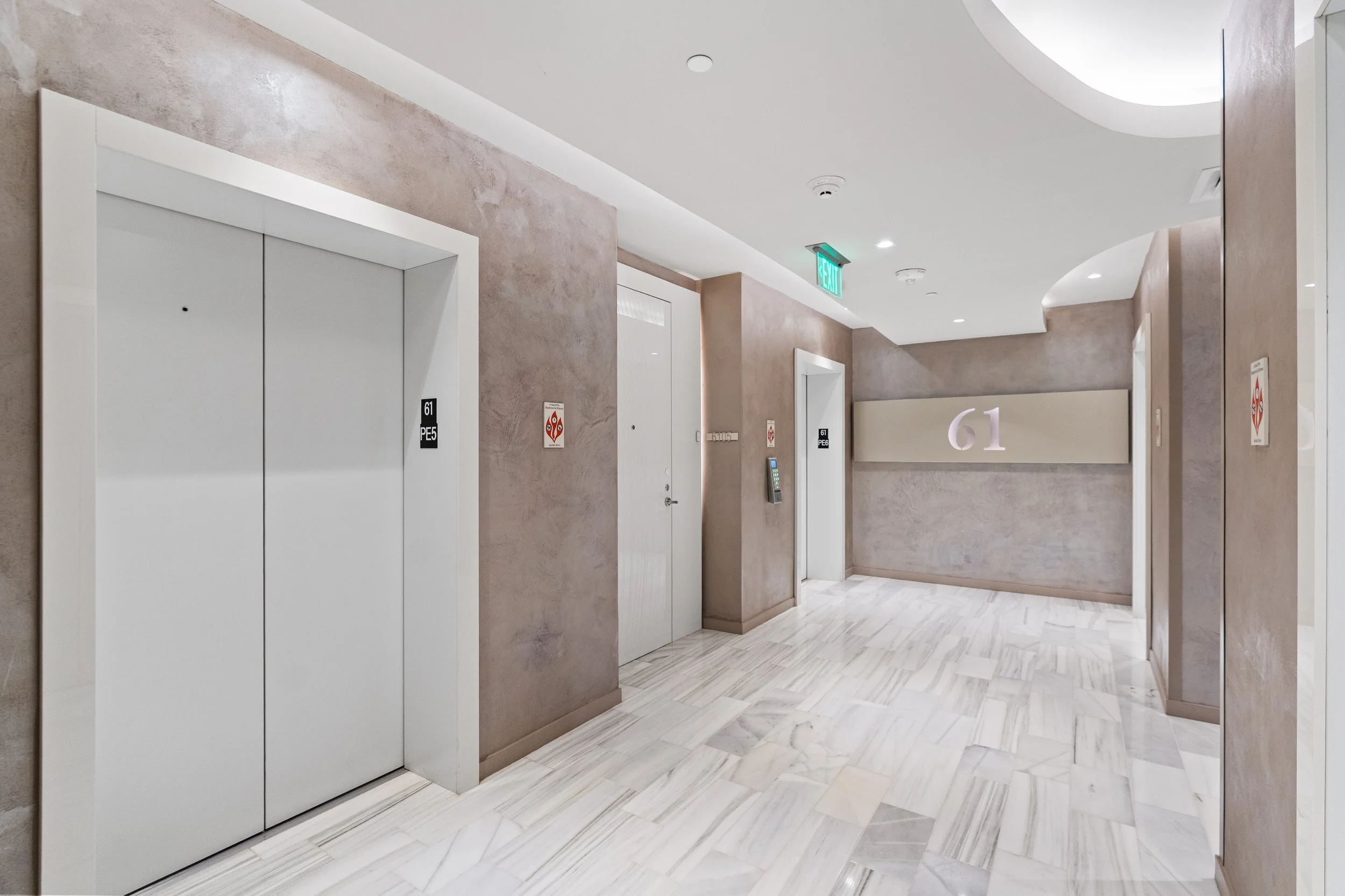 An indoor hallway with beige textured walls, white ceiling with recessed lighting, and an elevator with white doors. There is a digital keypad next to the elevator, a large illuminated number 61 on a panel, and an illuminated green exit sign. The flo