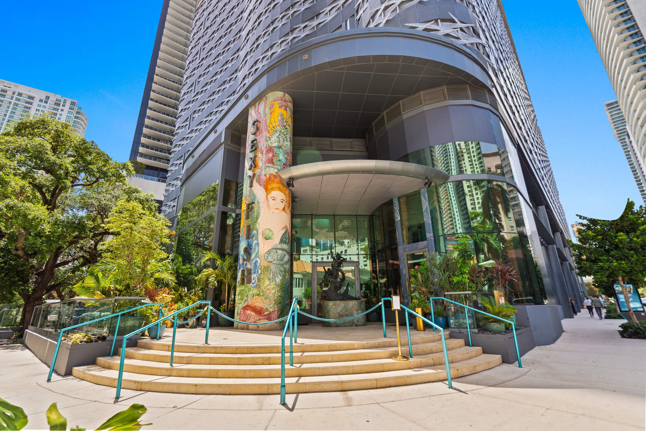 View of a modern tall building with a curved entrance and steps, surrounded by greenery and trees on a sunny day.