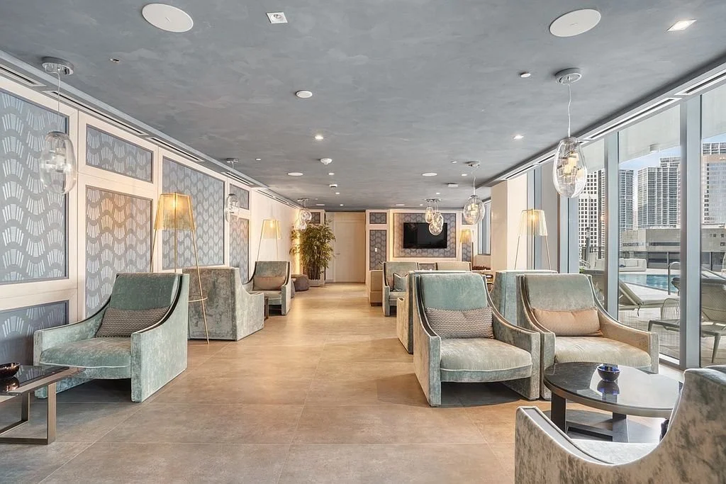 A spacious, modern lounge area with plush light teal armchairs, side tables, and floor lamps. Large windows reveal a cityscape with tall buildings, and a flat-screen TV is mounted on the far wall.