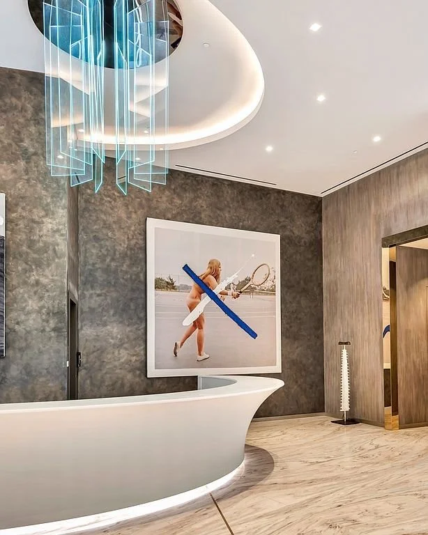 Modern hotel lobby with a white curved reception desk, textured grey wall, large framed photo of a woman with a tennis racket, and a unique blue chandelier hanging from the ceiling.