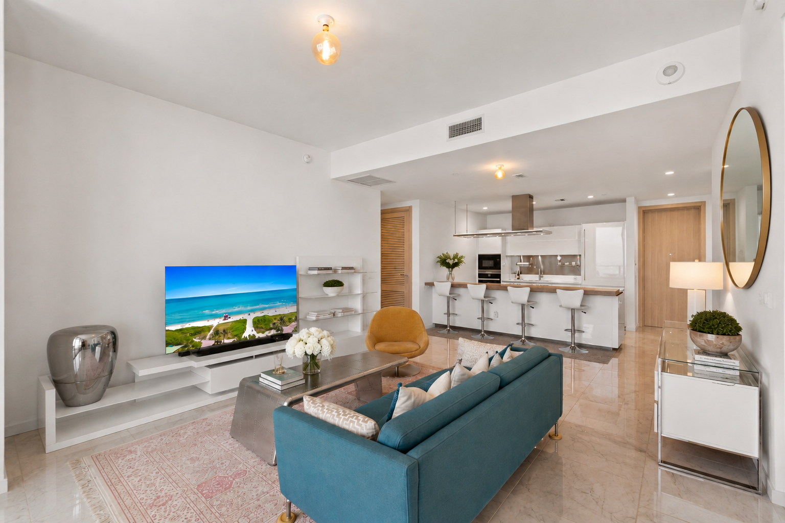 Open-concept living room and kitchen with modern decor, a blue sofa, a yellow chair, a TV displaying a beach scene, and white bar stools.