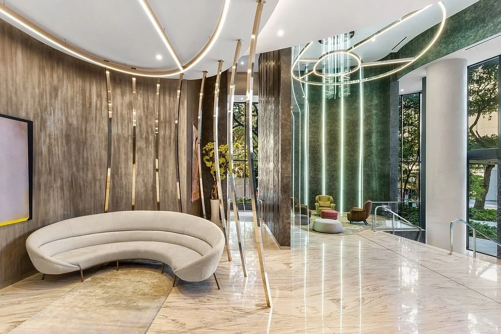 Modern hotel lobby with curved white sofa, marble floor, and decorative strip lighting, featuring seating areas near large windows with trees outside.
