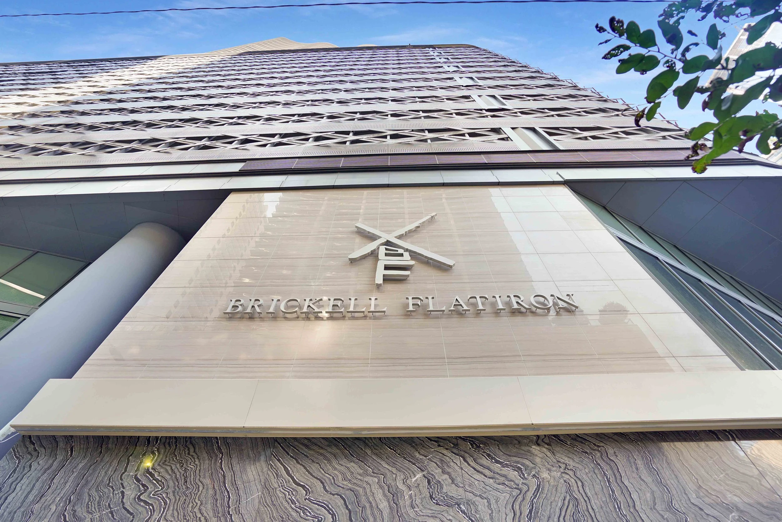 Upward view of the Brickell Flatiron building with a modern architectural facade, a large column on the bottom left, and a sign displaying the building's name on the front. A portion of a tree is visible on the right side, and the sky is clear blue.