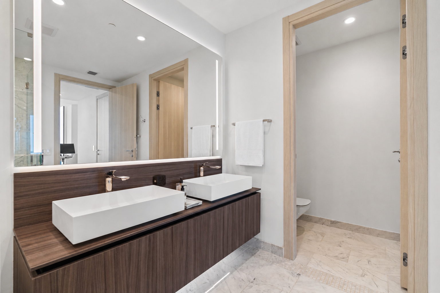 Modern bathroom with a double vanity featuring rectangular white sinks on a dark wood countertop, large mirror, towel racks with white towels, and a separate toilet area visible through an open doorway.