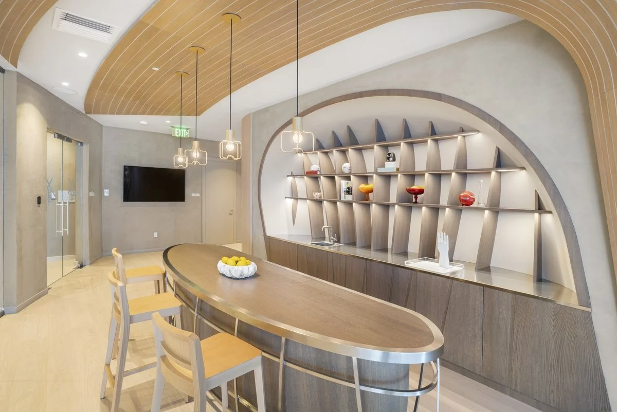 Modern room with a large wooden oval table, four chairs, a decorative wall shelf with colorful bowls, a flat-screen TV, and wooden ceiling accents.