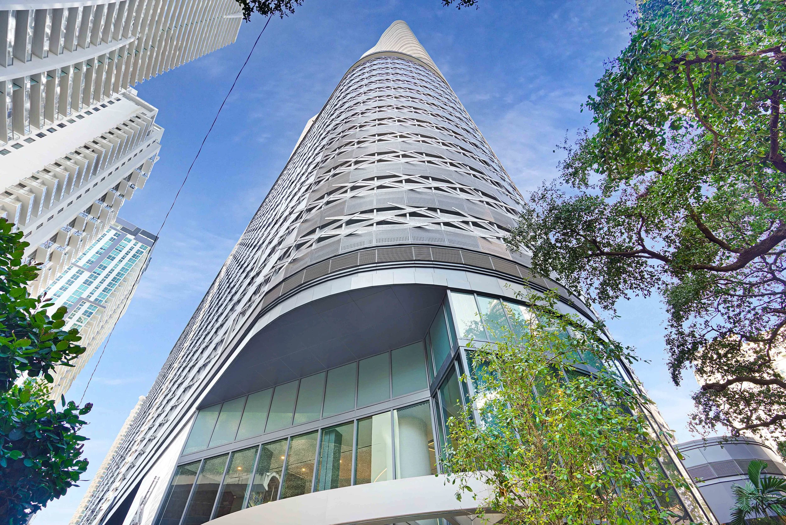 Low-angle view of a modern skyscraper with a curved, patterned facade, surrounded by green trees against a blue sky.