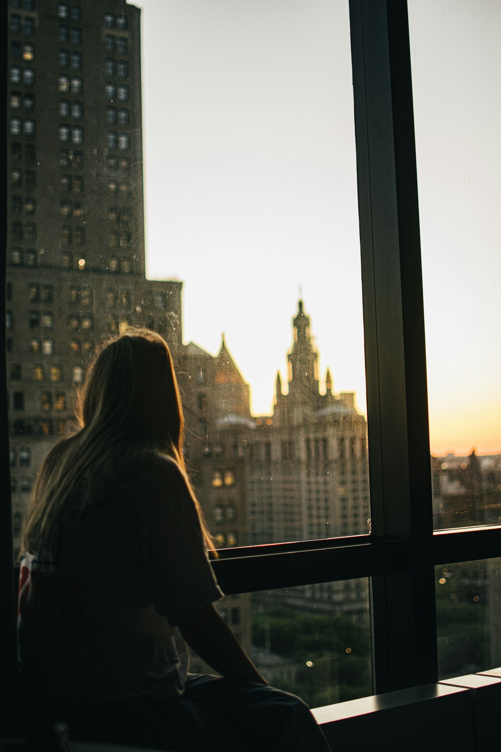 Woman sitting by a large window overlooking city buildings at sunset