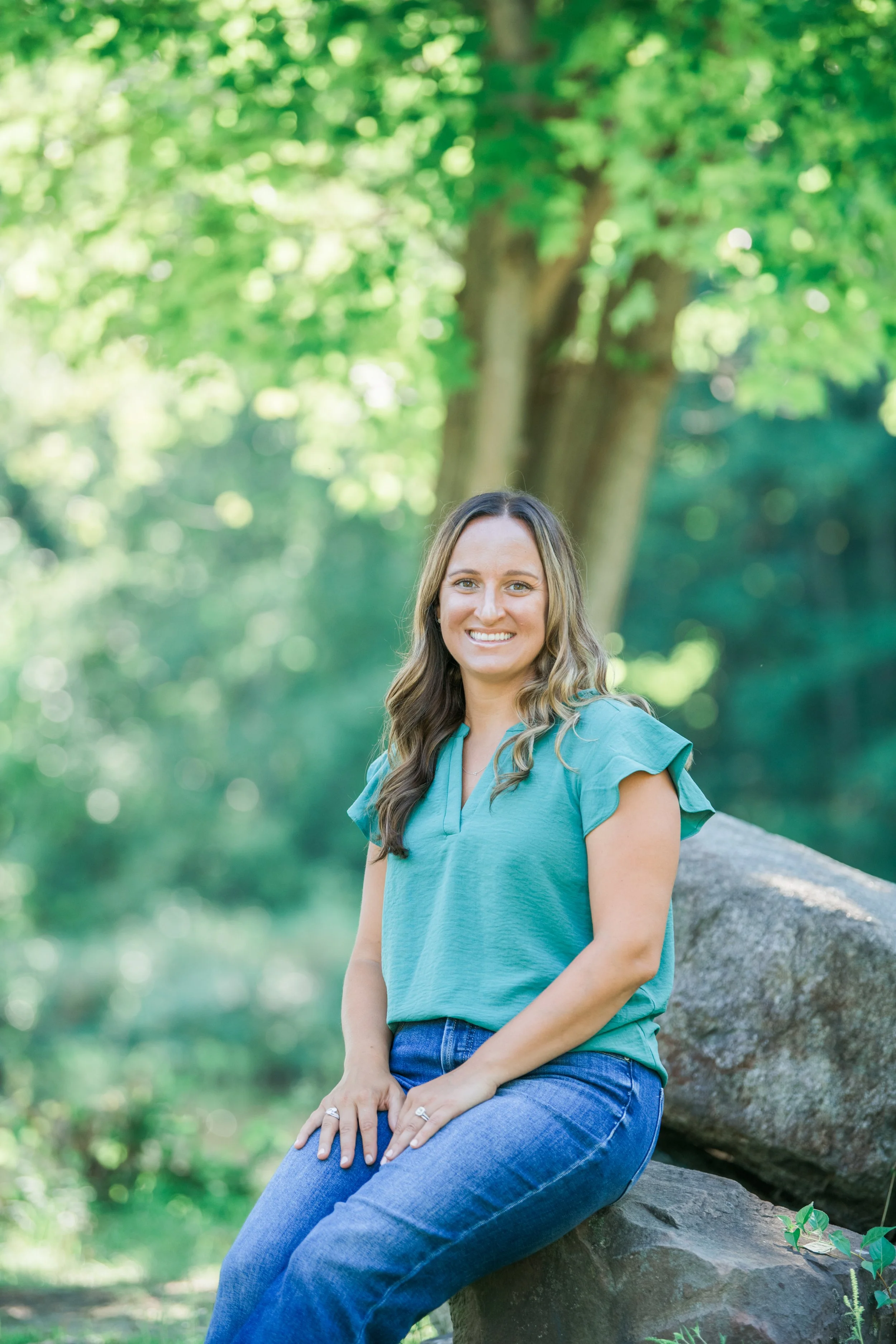 Melissa Parrish, LPC, sitting on a rock and smiling, with a blurred forest background