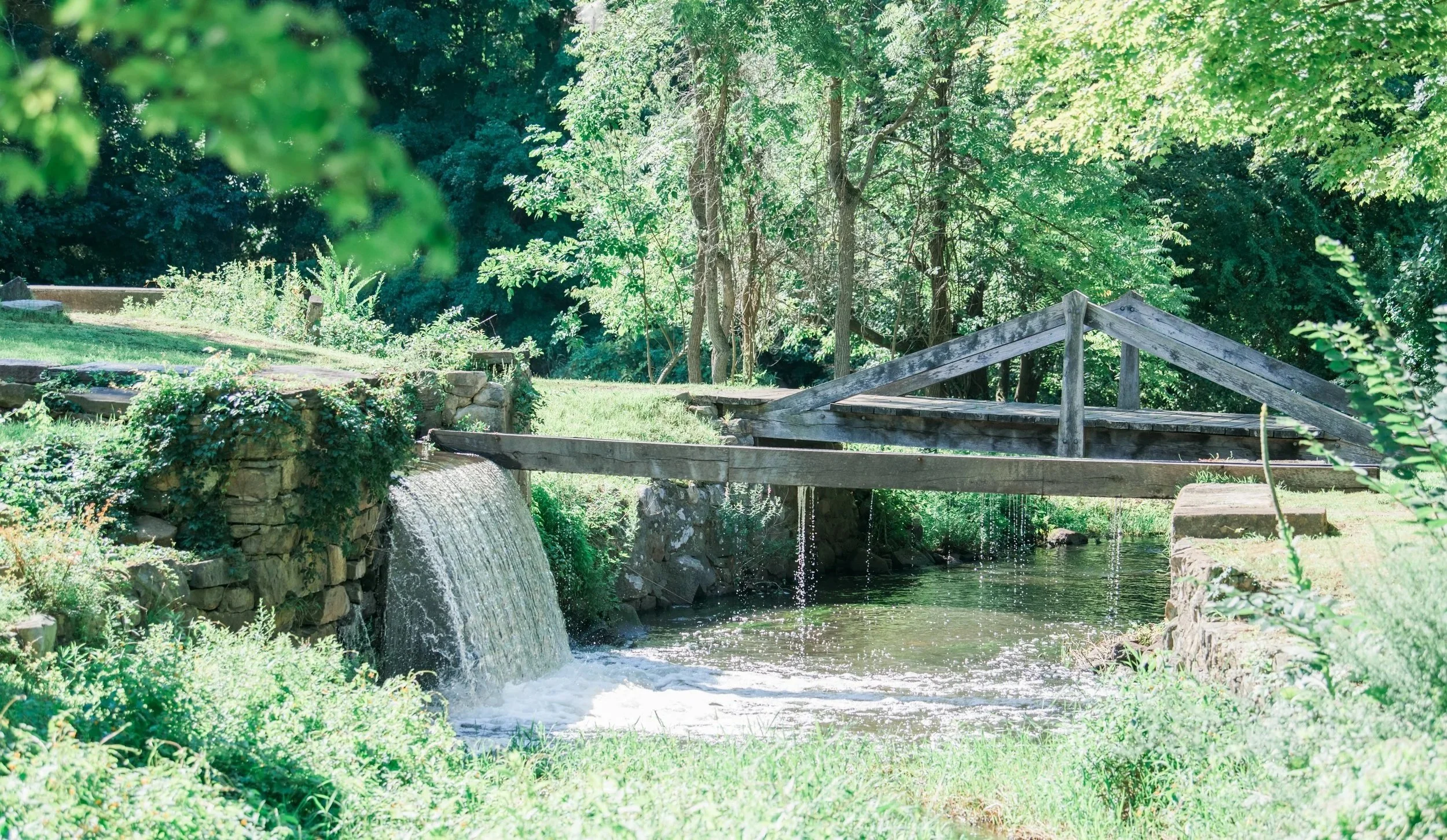 Small waterfall beside a wooden bridge and stone wall surrounded by trees