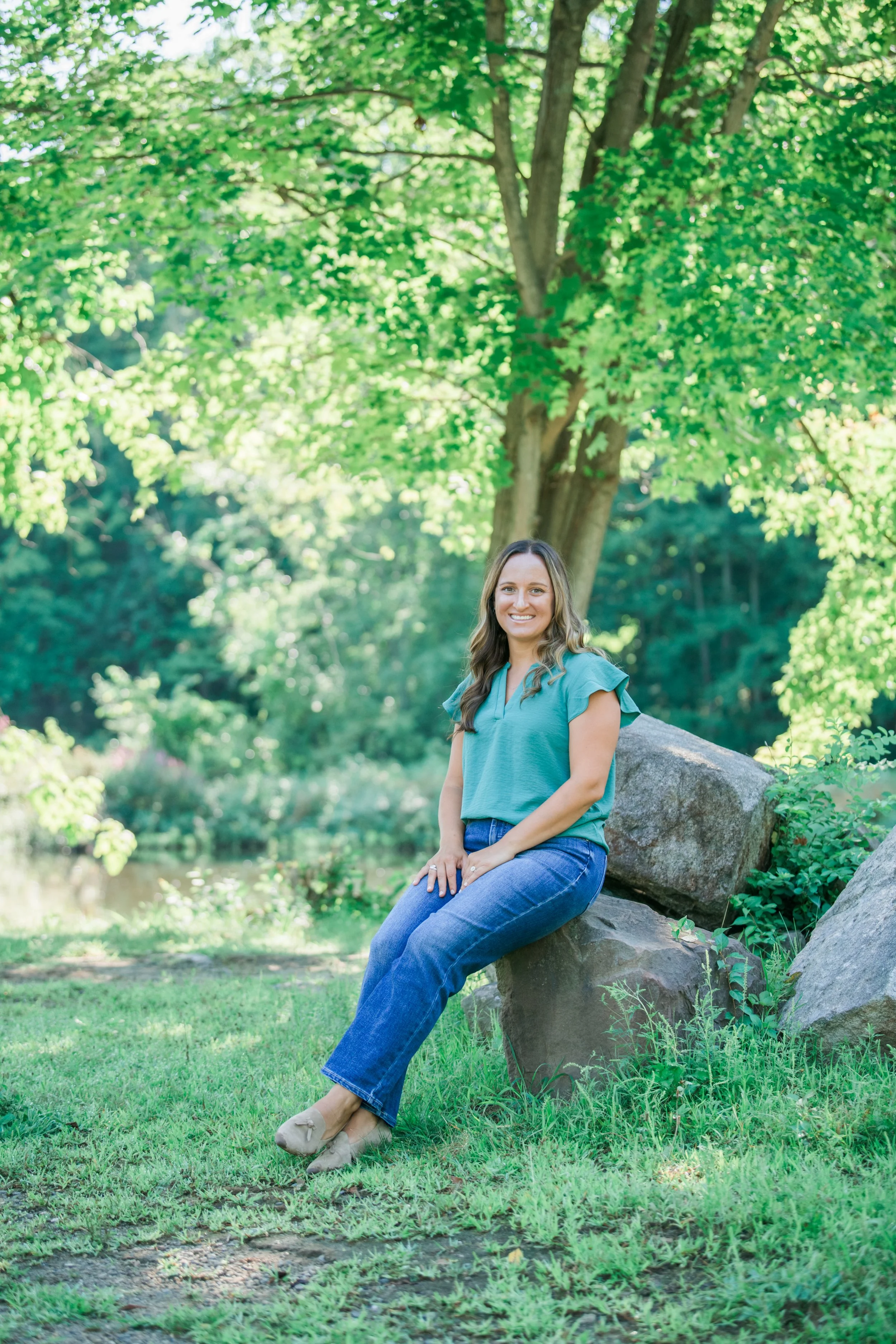 Melissa Parrish, LPC, sitting on a rock in a full-body shot with trees and water behind her