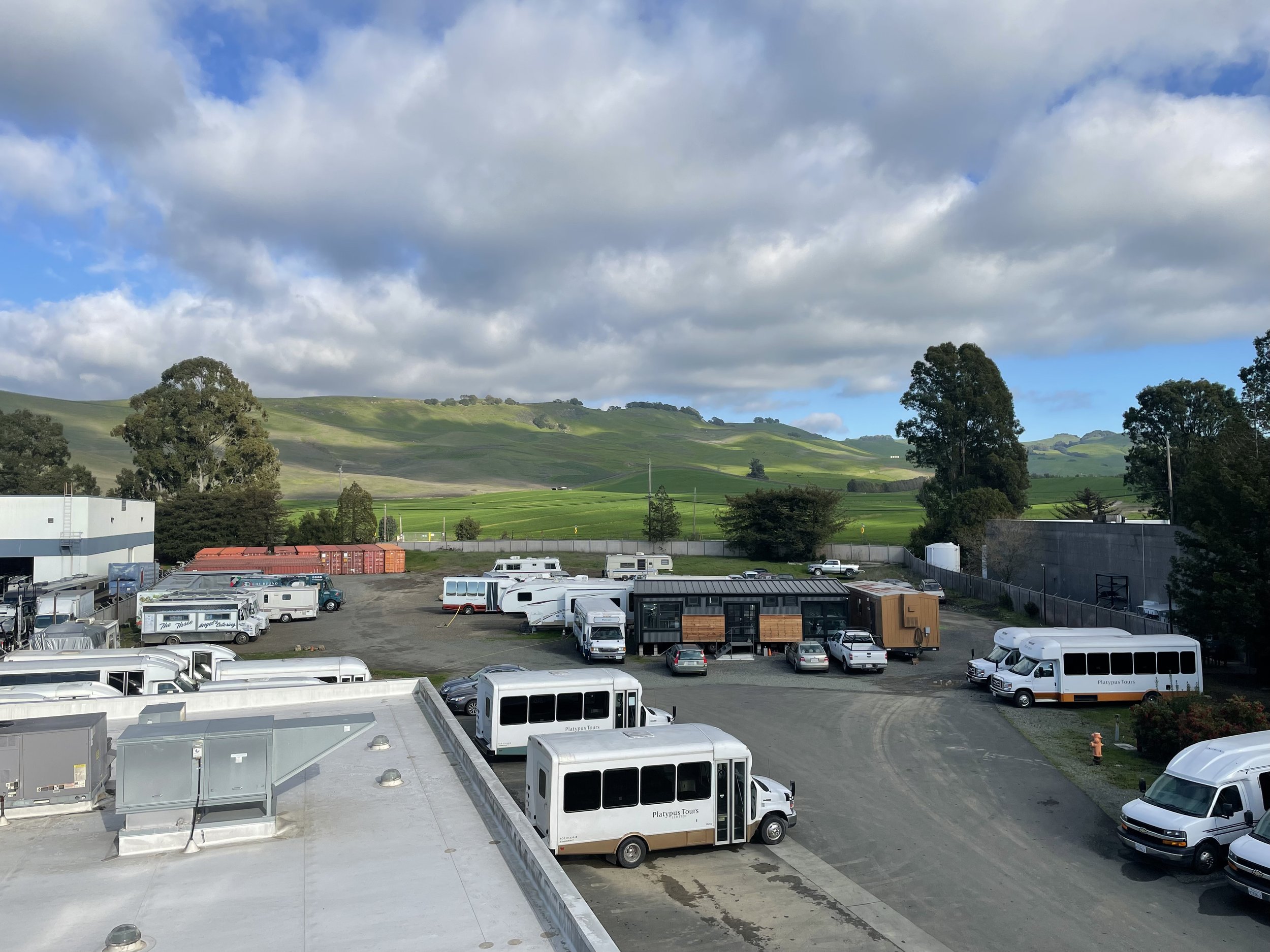 Parking lot with numerous recreational vehicles and trailers, surrounded by trees and green rolling hills under a partly cloudy sky.