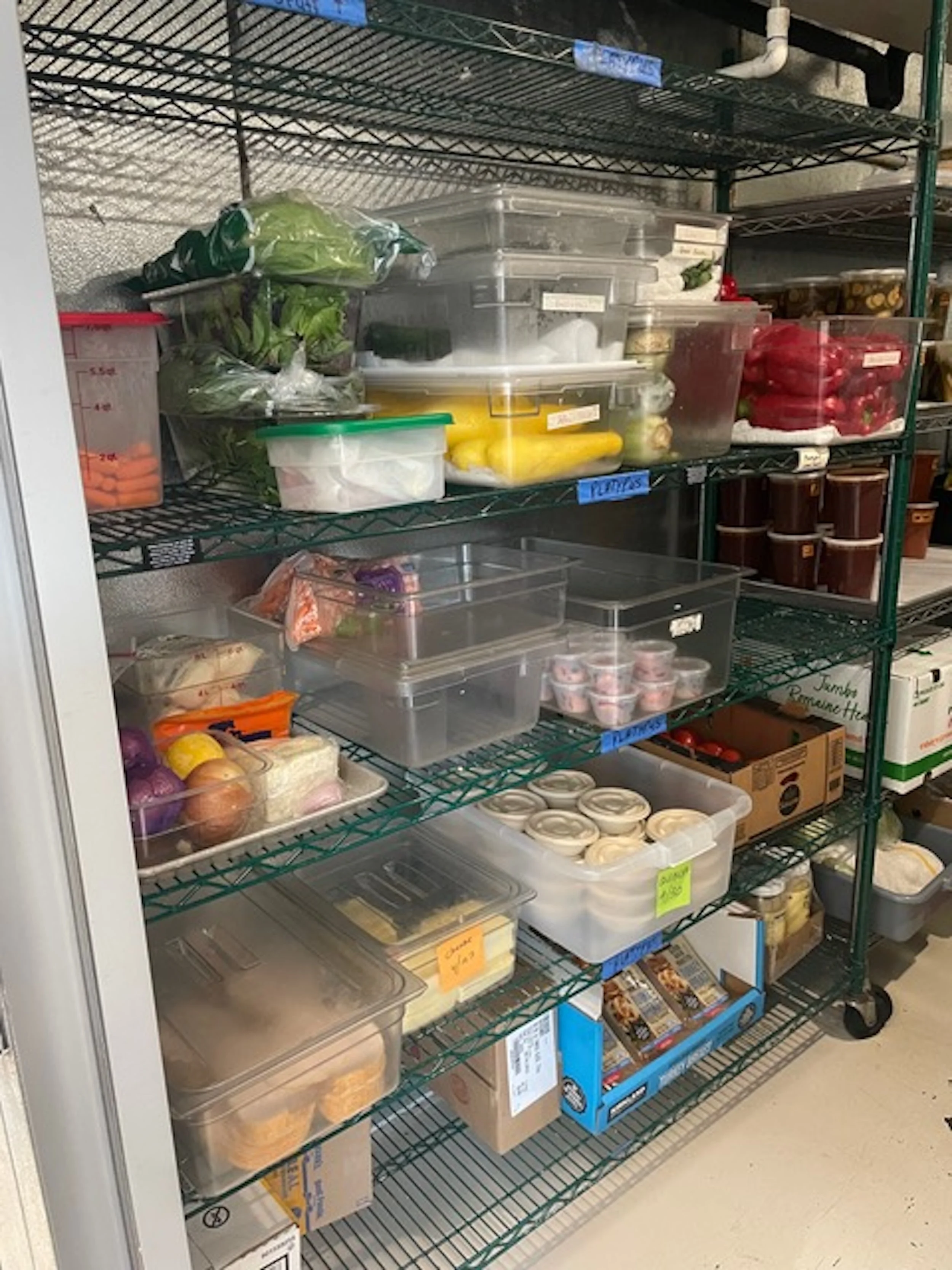 Metal storage shelves filled with various food items in clear plastic containers and boxes, including vegetables, yogurt, and packaged goods.