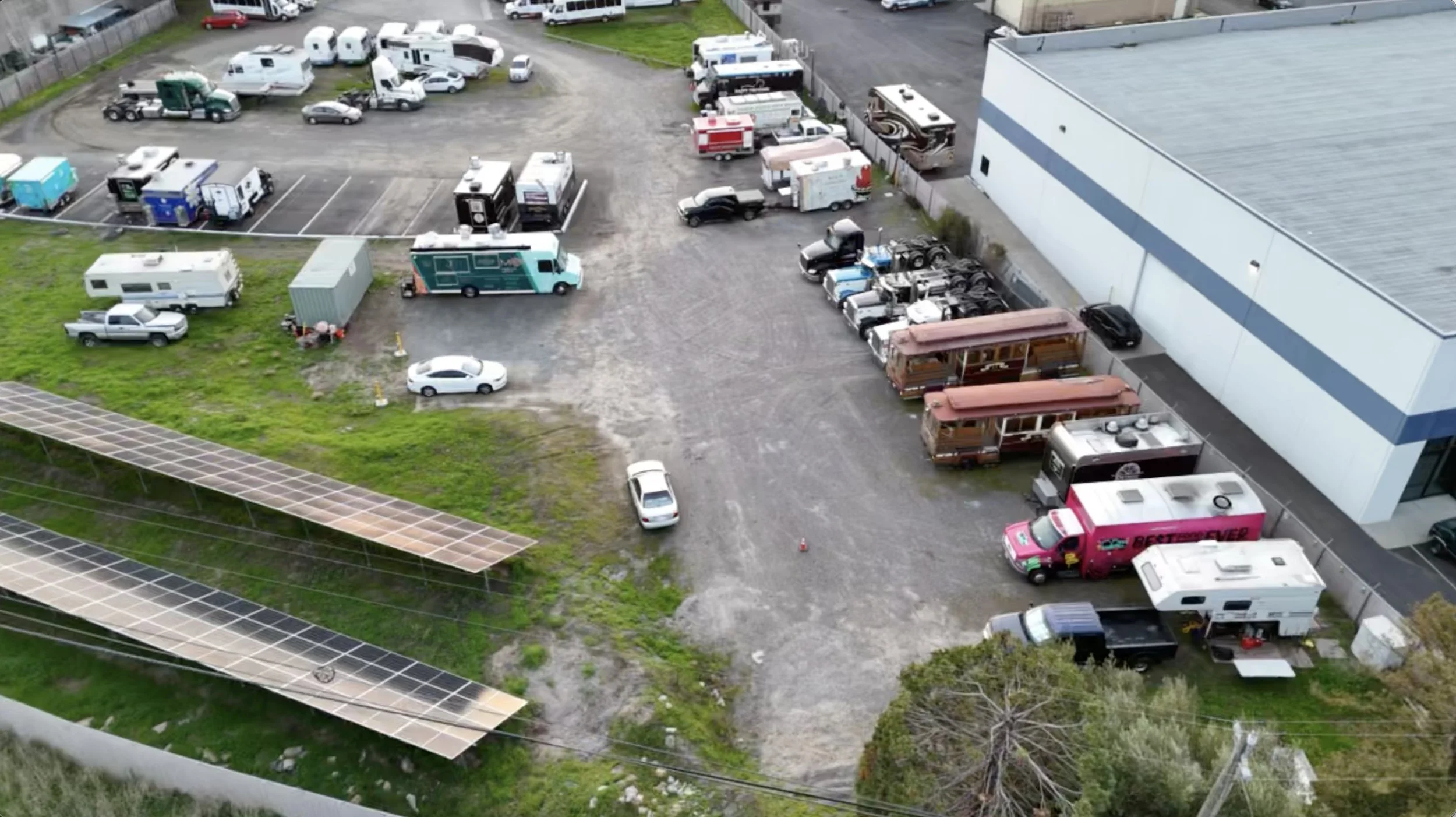 An aerial view of a parking lot filled with various vehicles, including RVs, trucks, trailers, and cars, adjacent to a large warehouse or industrial building and green grassy areas.