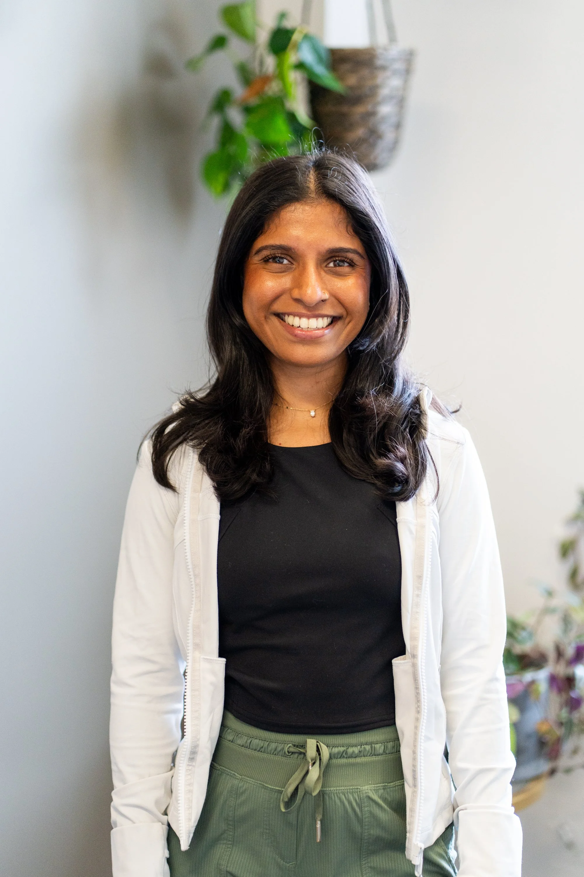 A young woman with long dark hair, smiling, standing indoors against a light-colored wall with a potted plant in the background.