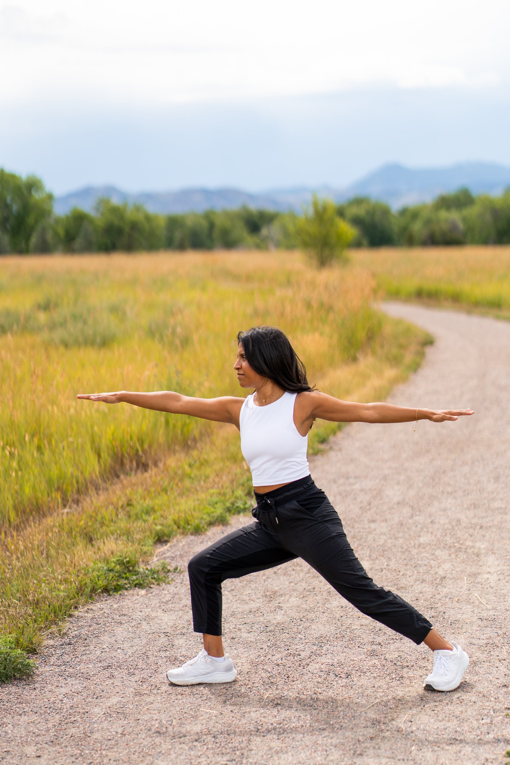 A woman practicing yoga outdoors on a dirt path in a natural setting, with her arms extended, wearing a white sleeveless top, black pants, and white sneakers.