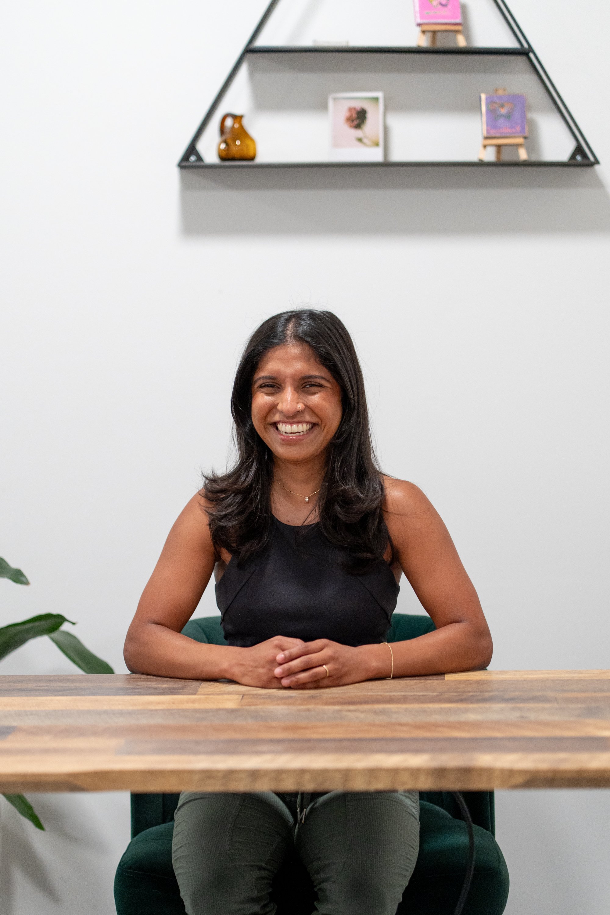 A woman with long black hair sitting at a wooden table, smiling, wearing a black sleeveless top and jewelry, with a white wall and decorative shelves in the background.