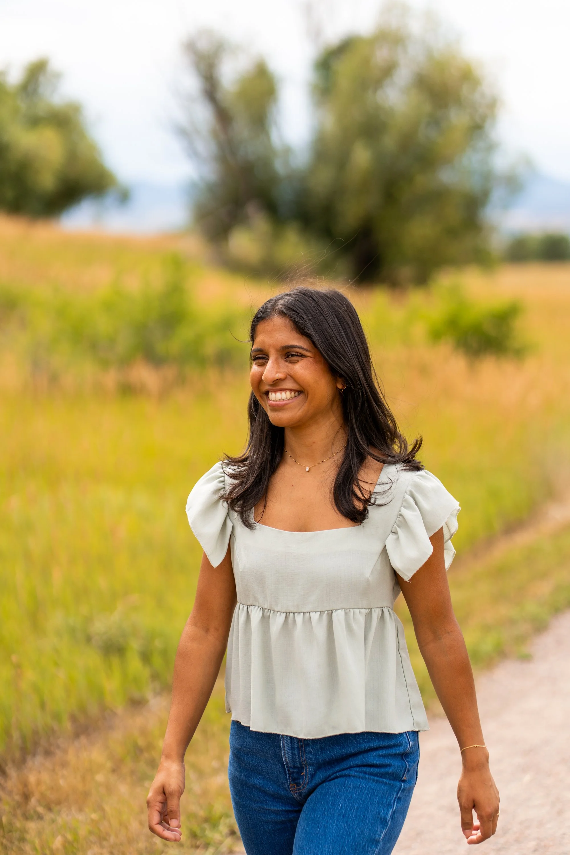 A woman with long dark hair smiling and walking outdoors in a field with greenery and trees in the background.