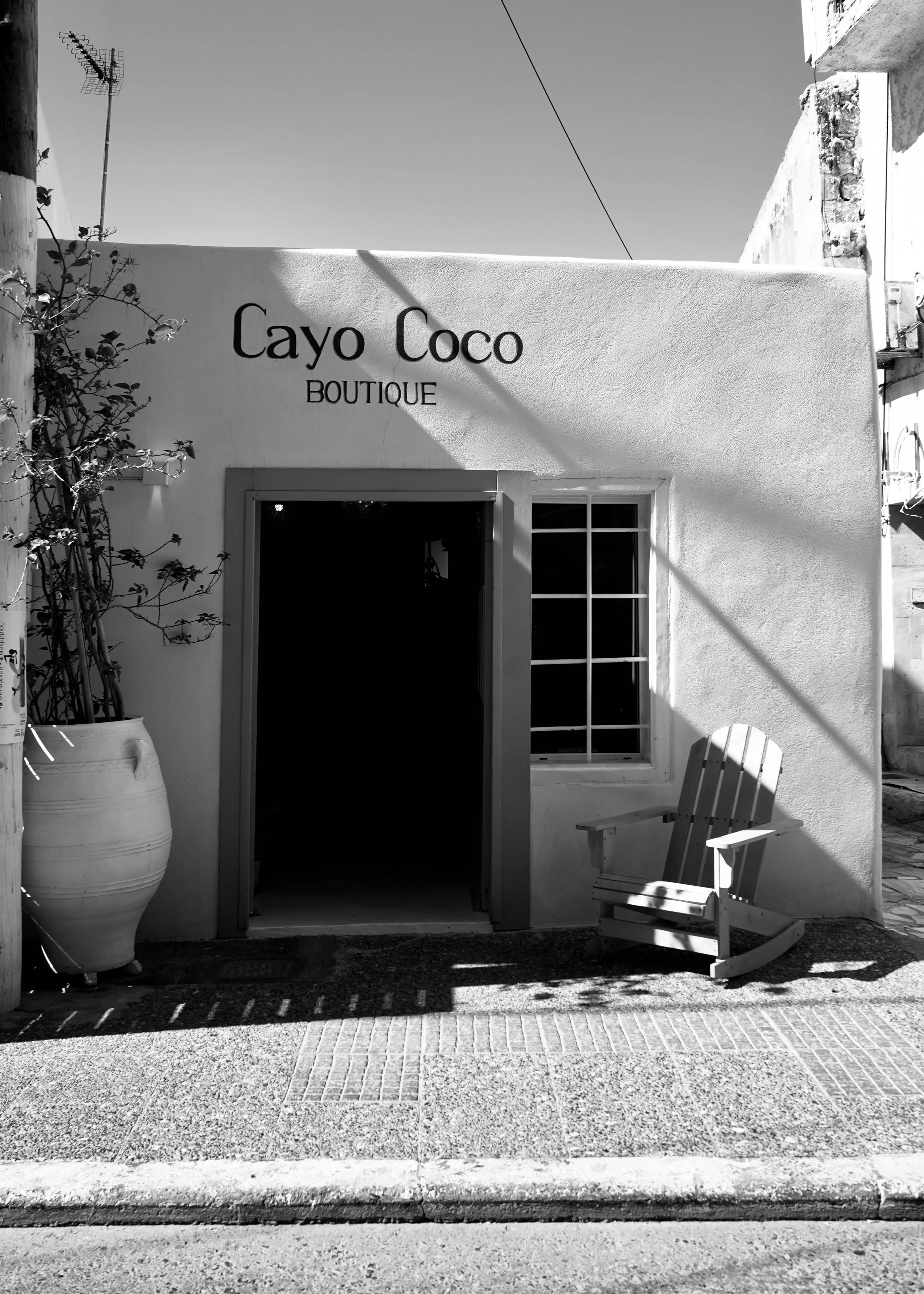 En hvid væg med skiltet 'Cayo Coco BOUTIQUE'. Der er en stor keramisk plantekrukke med en vækst af planter til venstre, og en hvid træstol på højre side foran væggen. Der er også en åben dør med et vindue ved siden af, og skygger falder på jorden.