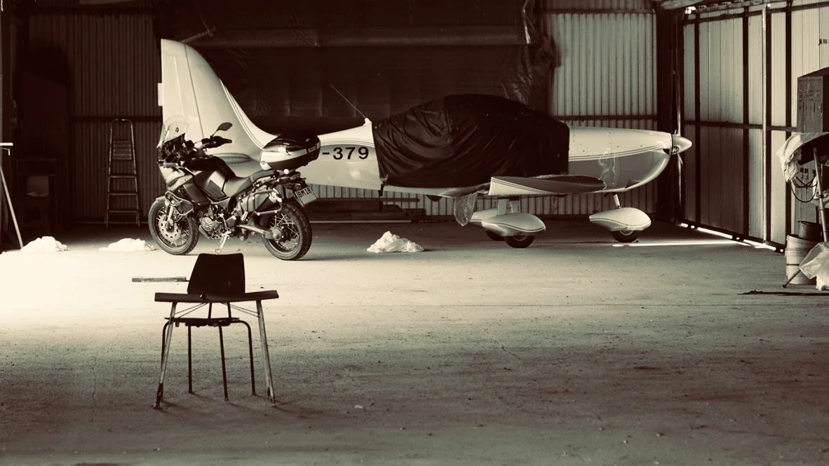 En flyvemaskine, der er dækket delvist til, og en motorcykel, der står ved siden af, i en hangar eller lagerbygning. Der er en tom stol foran, og nogle ting i baggrunden.