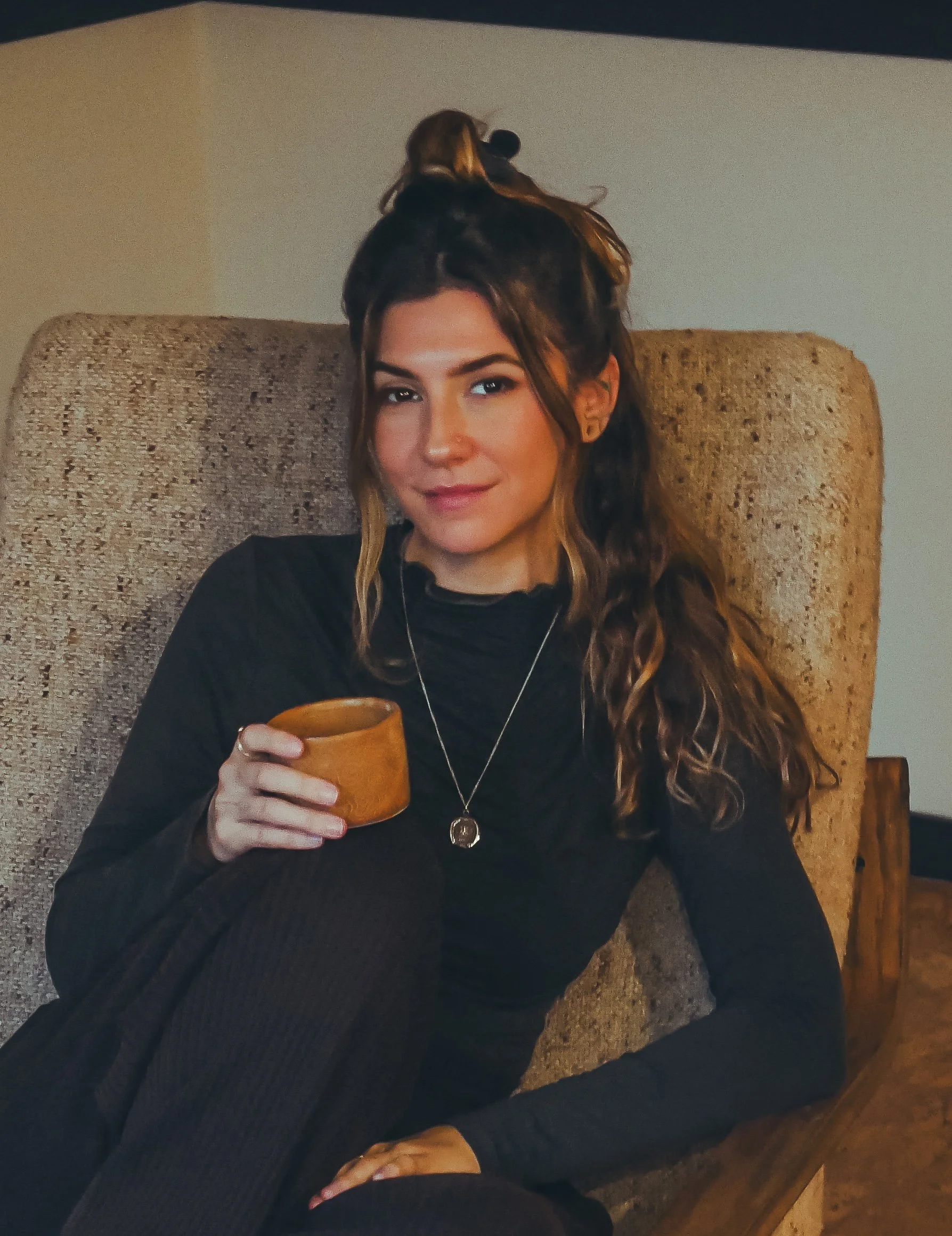 A young woman with long wavy hair sitting on a beige cushioned armchair, holding a small orange terracotta cup, wearing a black long-sleeve top and a necklace with a circular pendant, in a cozy indoor setting.