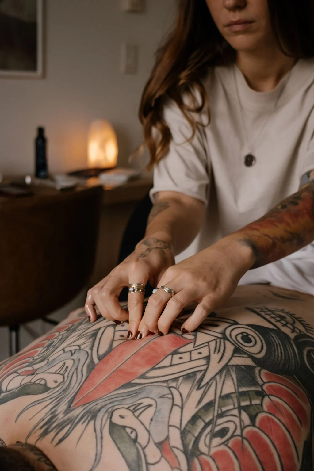 A man with tattoos on his back receives acupressure from a woman in a white tshirt. The background shows a dimly lit room with a lamp, a bottle, and books.