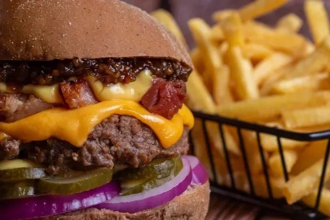 Close-up of a cheeseburger with pickles, onions, bacon, cheese, and sauce, next to a basket of French fries