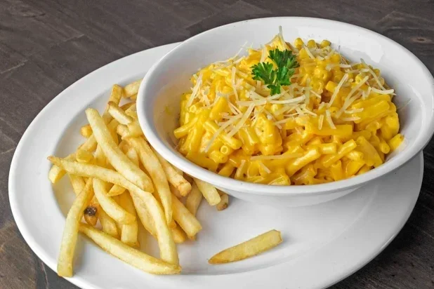 A white oval plate with French fries and a bowl of macaroni and cheese topped with shredded cheese and a parsley garnish.