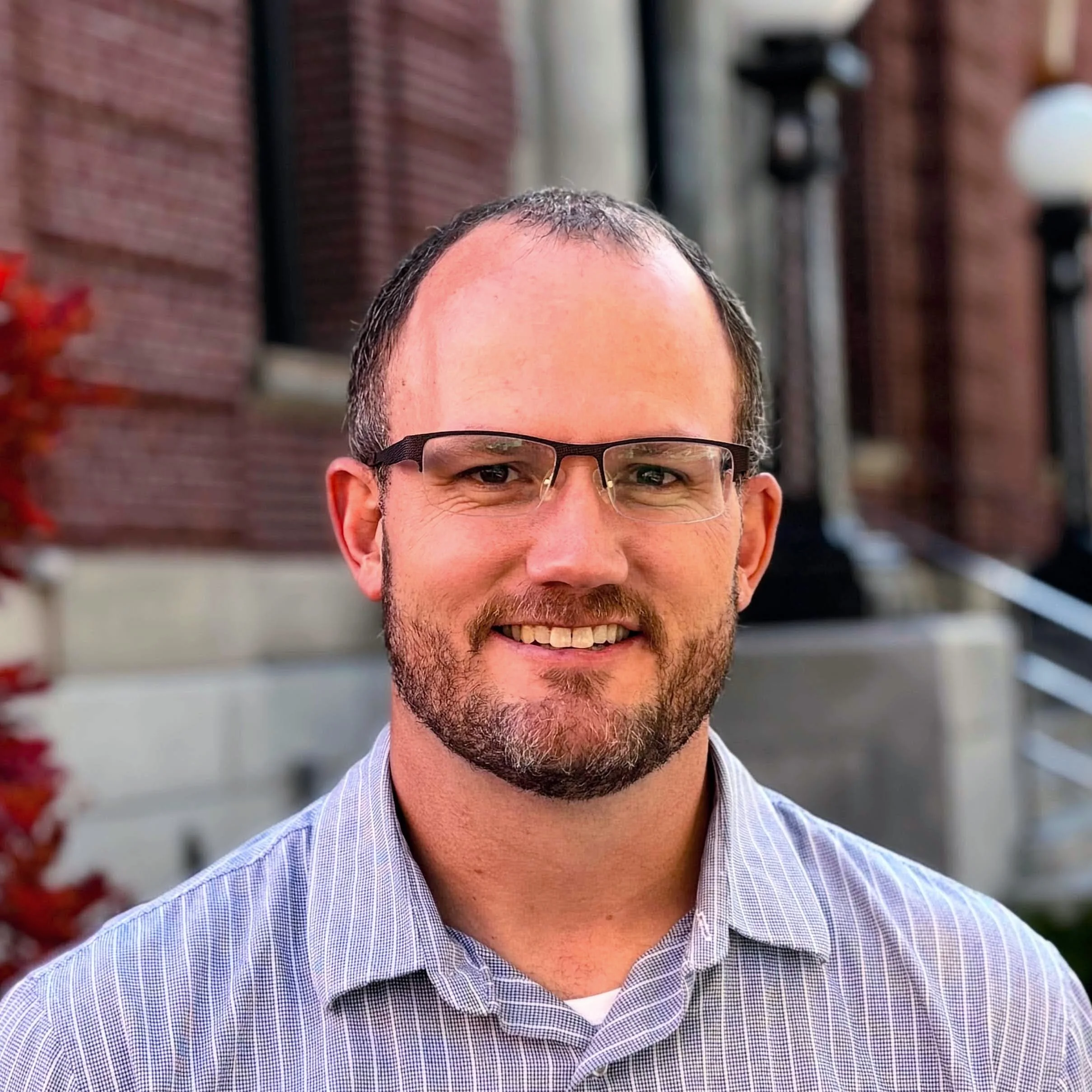 A smiling man with glasses and a beard wearing a checkered shirt, standing outdoors in front of a brick building with a window and some greenery.