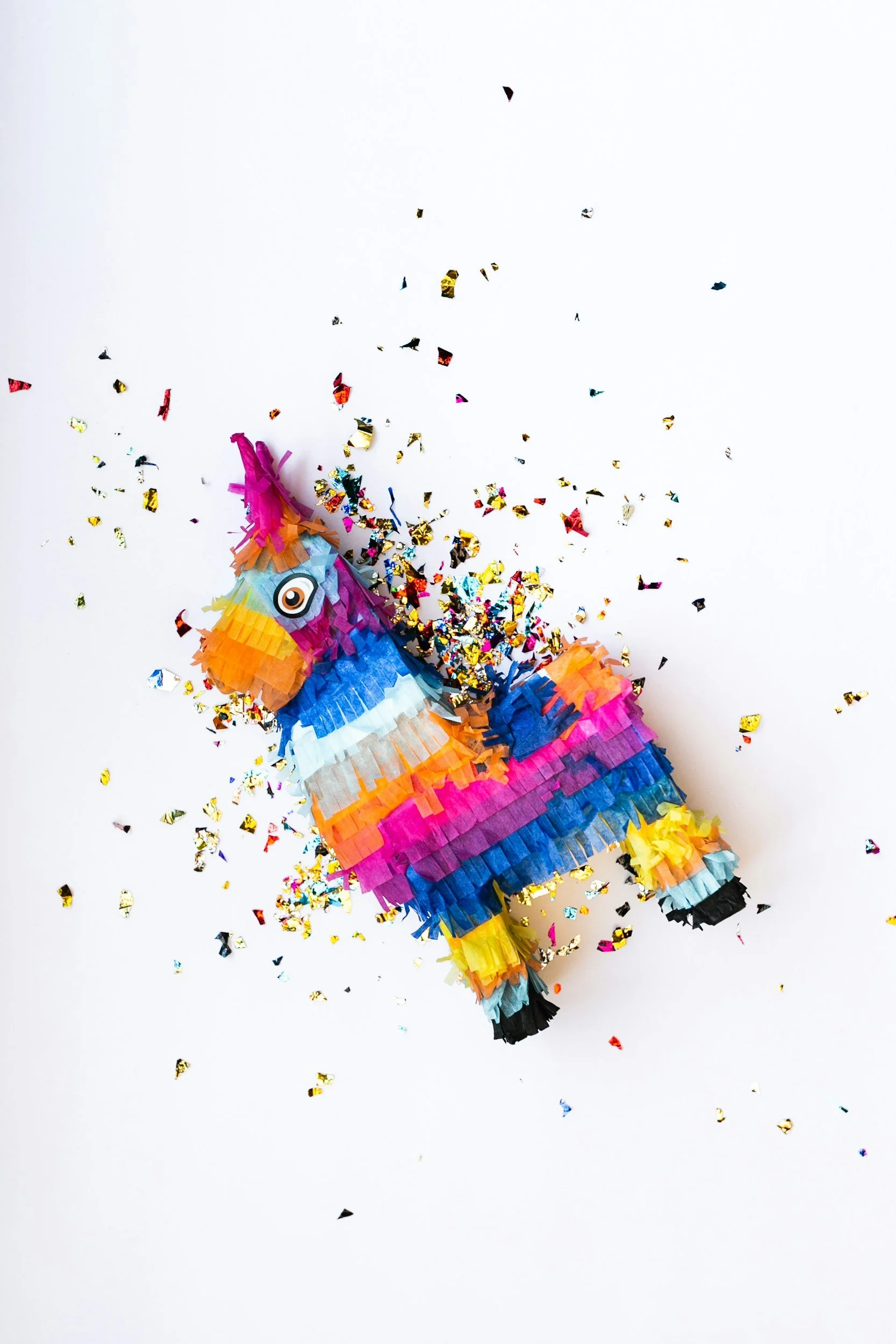 Colorful piñata shaped like a unicorn with a white horn, pink mane, and multicolored body, surrounded by scattered gold, red, and blue confetti on a white background.