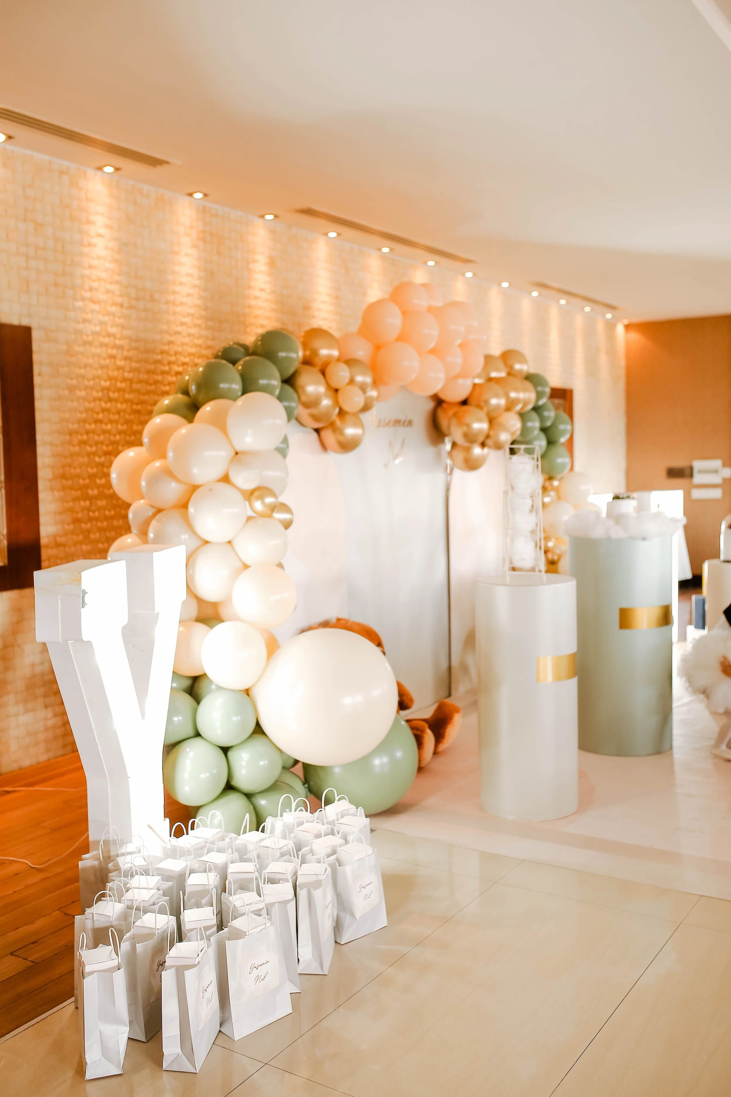 Decorative balloon arch in pastel colors, with white gift bags and white boxes with handles at a celebration or event, against a textured beige wall and wooden floor.