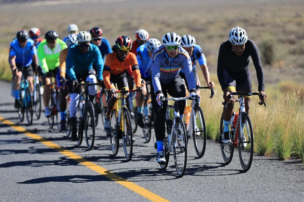 A group of cyclists riding on a paved road through a grassy landscape during the day, all wearing helmets and colorful cycling gear.