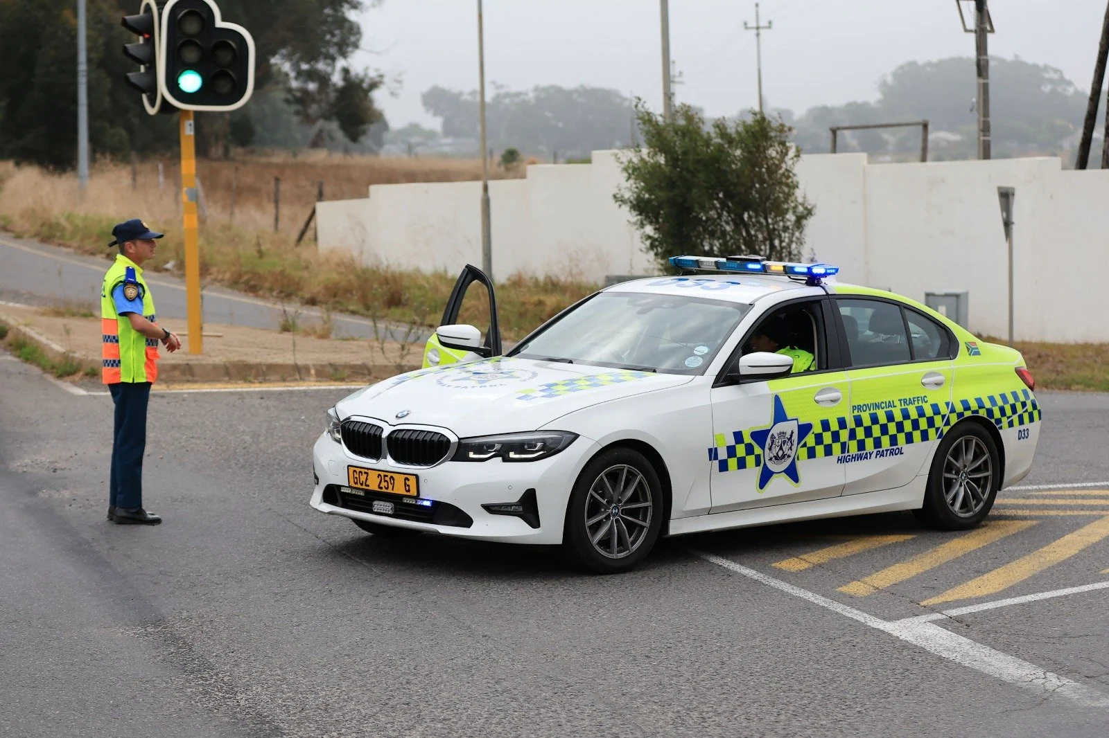 A police officer speaking to a driver inside a white marked police car on a road. The police car has blue and yellow markings and flashing lights on top. The officer is wearing a bright yellow reflective vest and a hat. The scene appears to be during