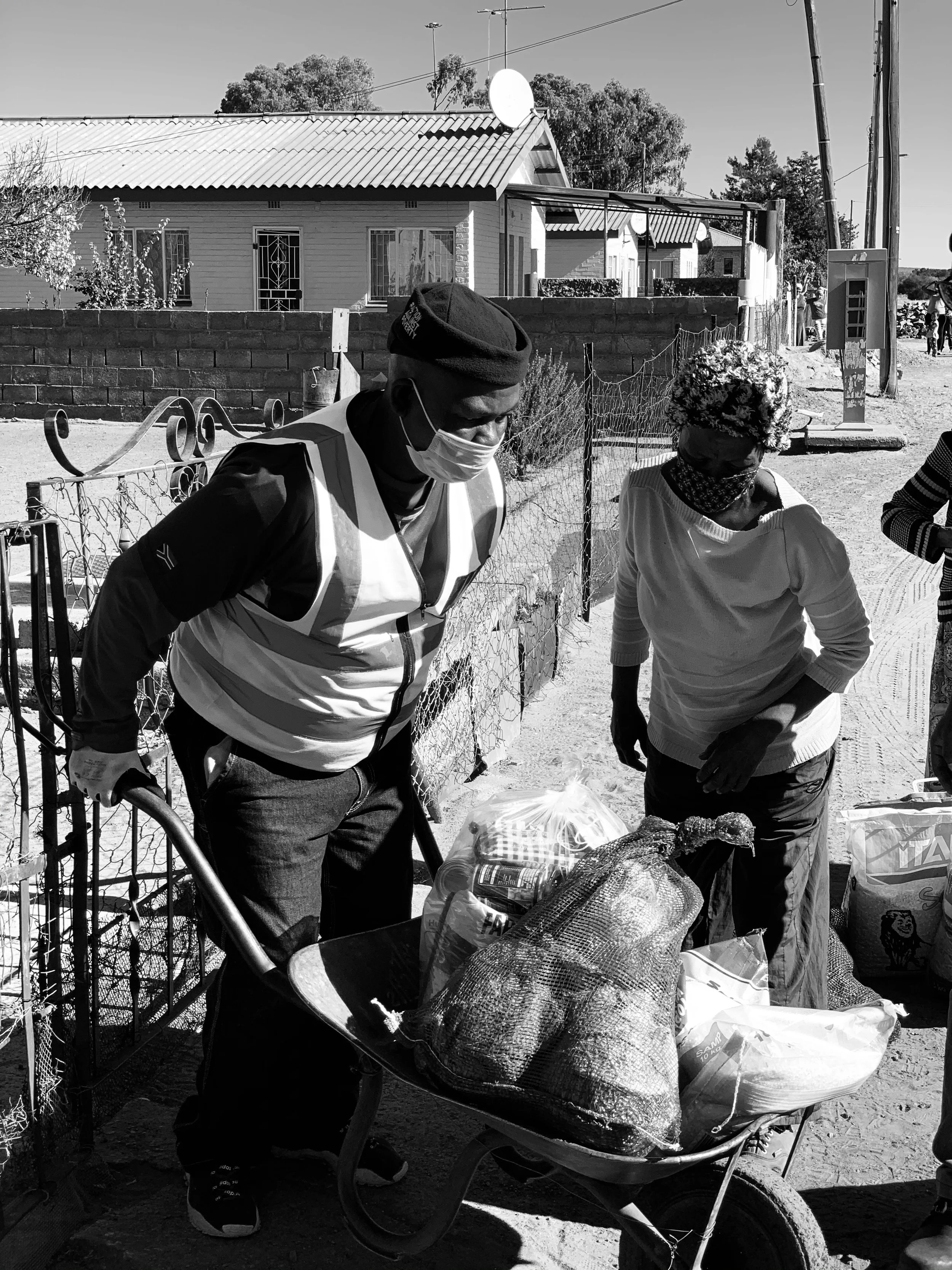Two people wearing masks and hats are getting groceries near a fence outside. One person is holding a shopping cart filled with bags, and there are houses and power lines in the background.
