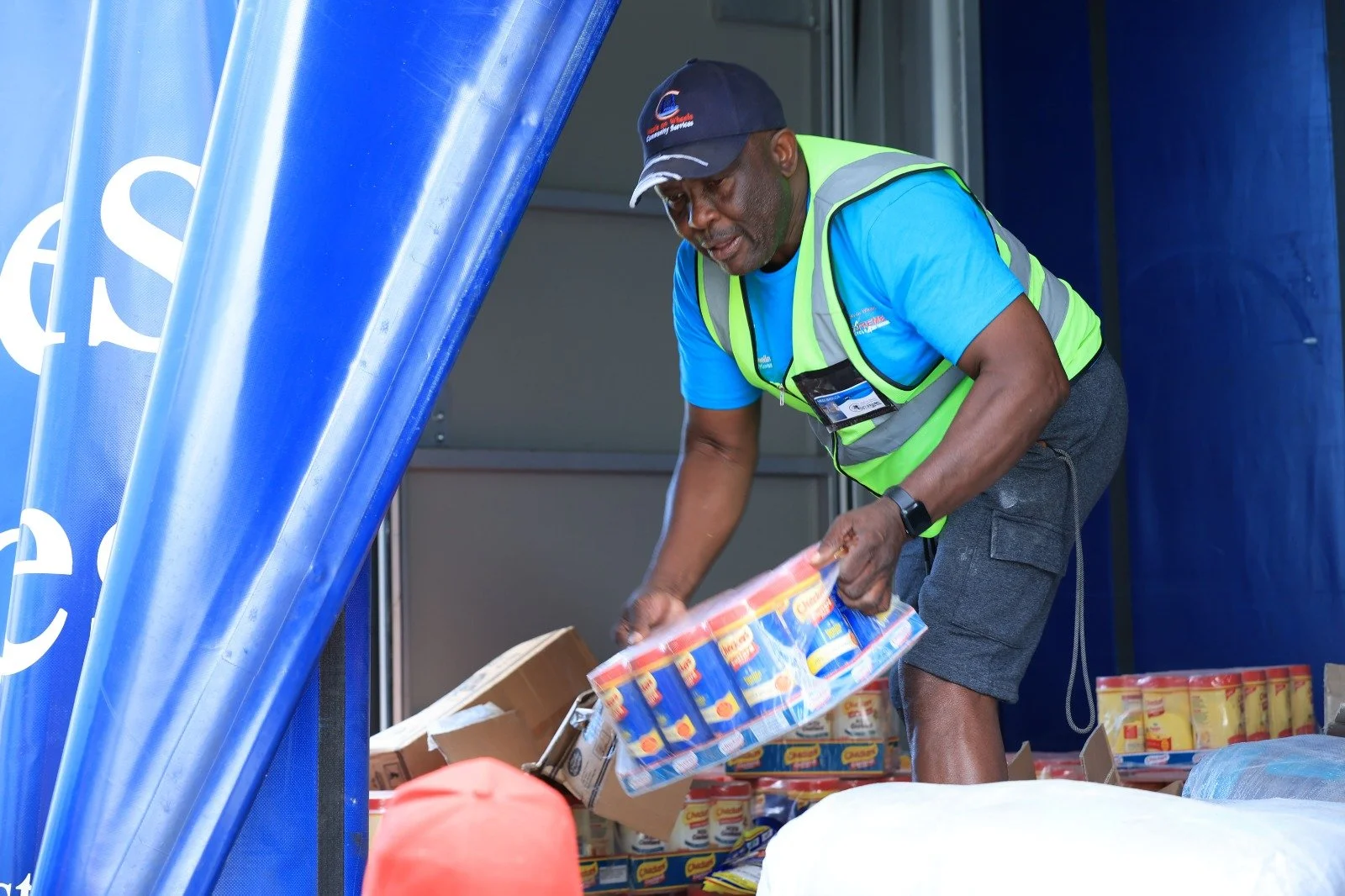 A man wearing a blue shirt, a green safety vest, and a cap is sorting canned goods at a food distribution event. There are boxes and cans of food around him.