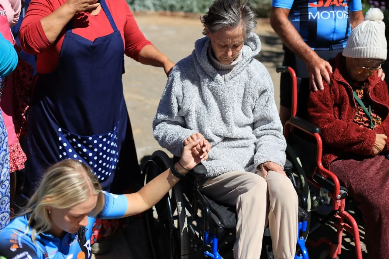 A group of people, including women in wheelchairs and a young girl, holding hands and offering support and comfort outdoors during daytime.