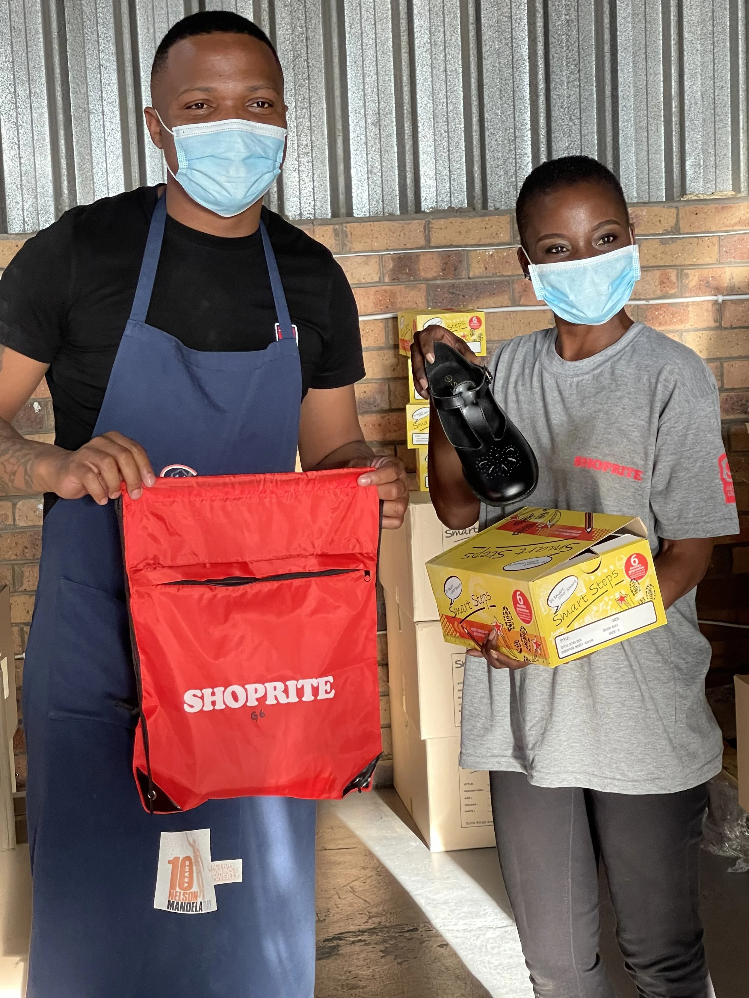 Two people wearing face masks, one holding a shoe and the other holding a box, standing in front of a brick wall with boxes stacked behind them. Shoe Drive with Shoprite