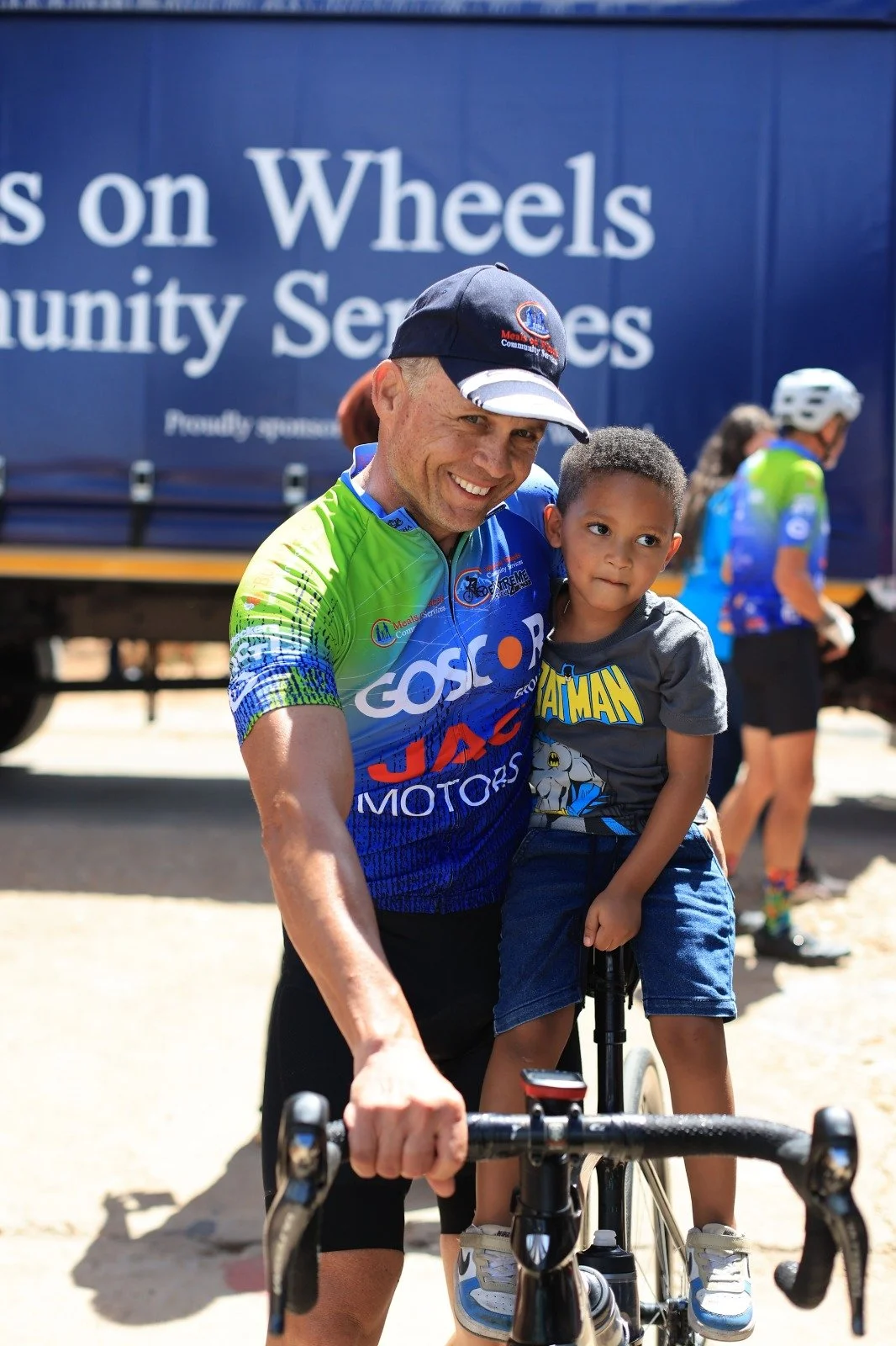 A man in a colorful cycling jersey is smiling and holding a young boy on a bicycle, both looking happy, with people and a large truck in the background.