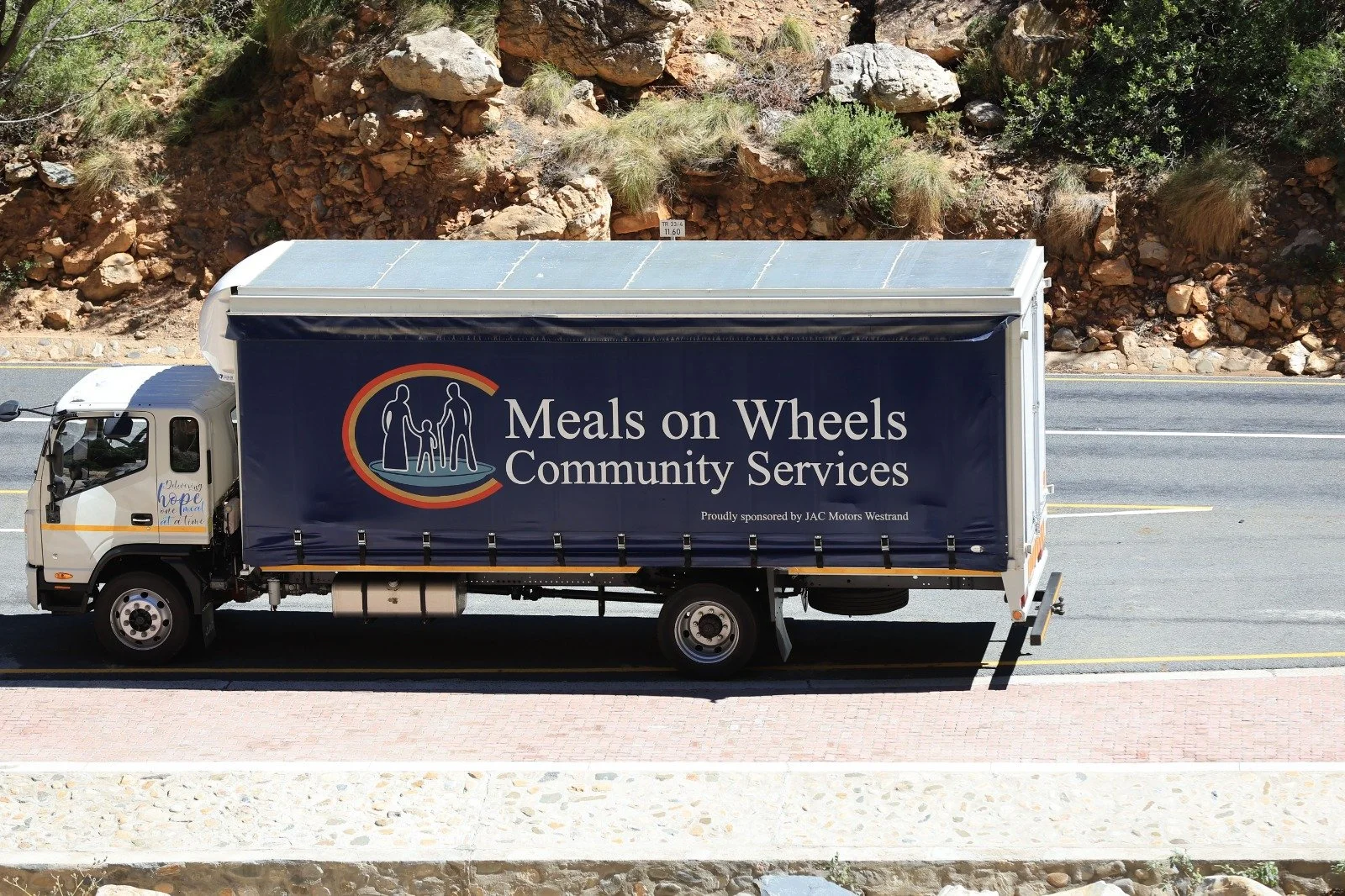 A truck with a large banner on its side that reads "Meals on Wheels Community Services" and features a logo of two adults and a child holding hands. The truck is driving on a road with a rocky hillside and some greenery in the background.