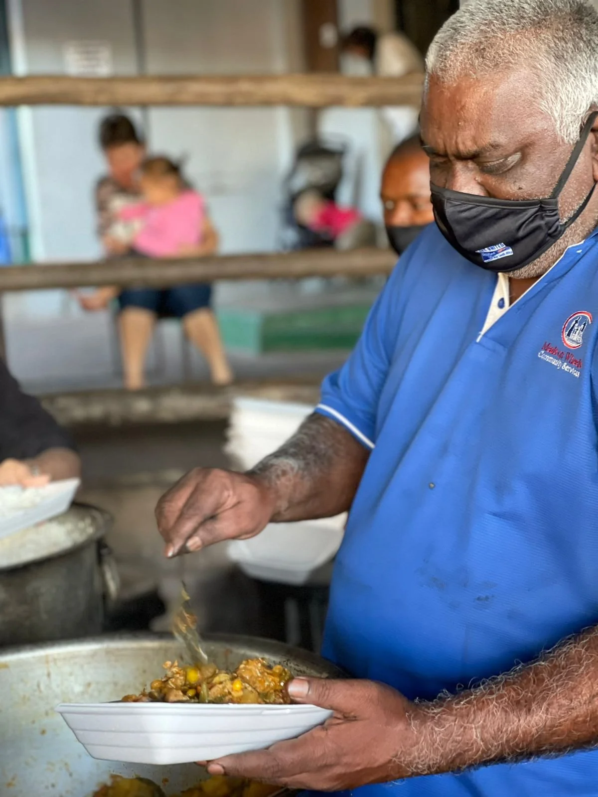 An older man with gray hair wearing a black face mask and a blue shirt is serving food from a large pot into a white bowl, with a blurred background showing a woman holding a child and other people. Caregiver Services
